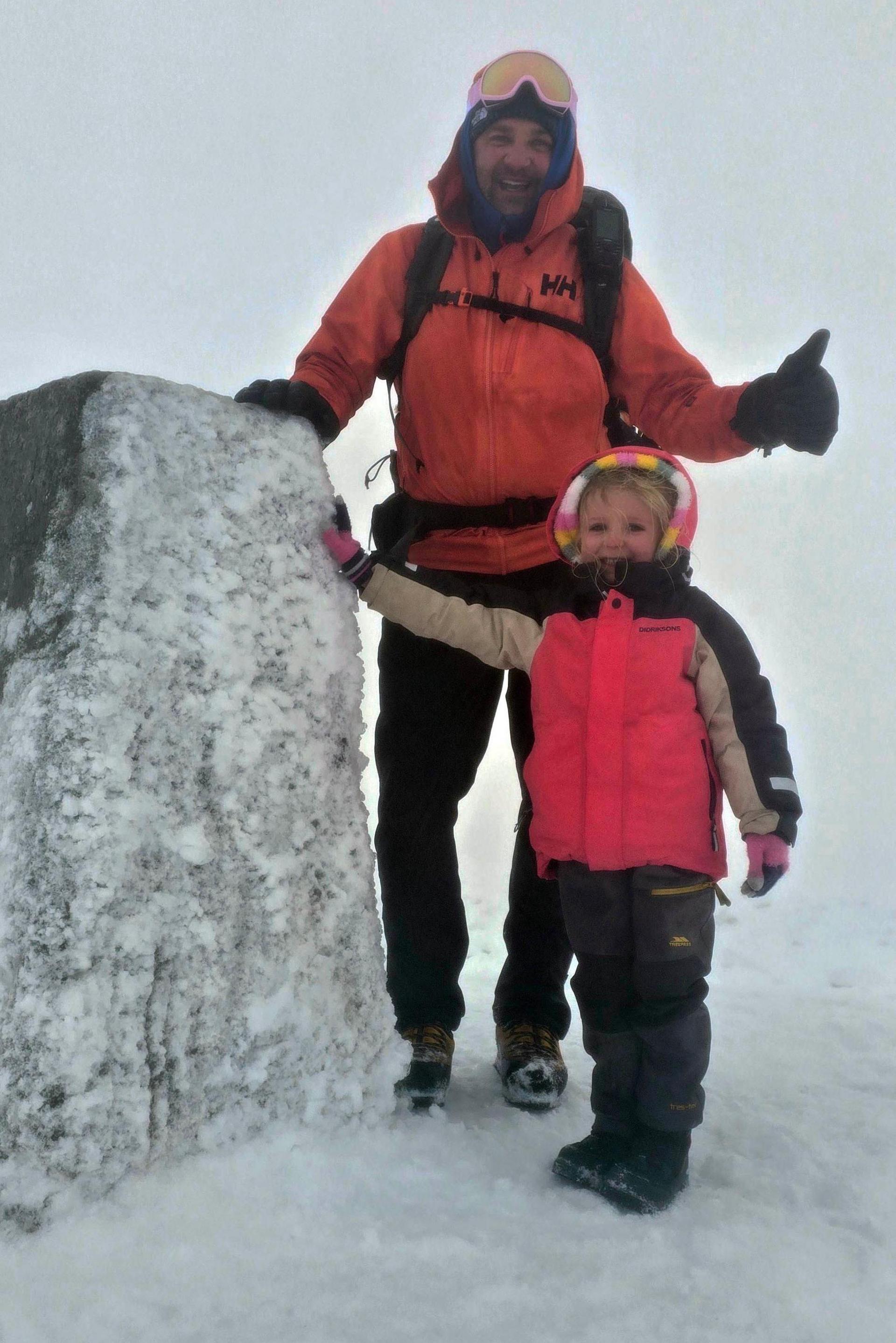Faye Moffat and her dad Craig smiling as they touch the concrete trig point on the summit of Ben Nevis which is covered in snow and ice. It is snowy and foggy. They are both wearing waterproof winter gear. Faye is in a bright pink jacket with beige and black sleeves. The hood is up over the top of the hood of a multi-coloured fleece. She is also wearing grey trousers, black boots and winter gloves. Craig is wearing an orange jacket with black trousers, gloves, and boots. He has ski goggles on the top of his head and a rucksack on his back. He is giving a thumbs-up sign.