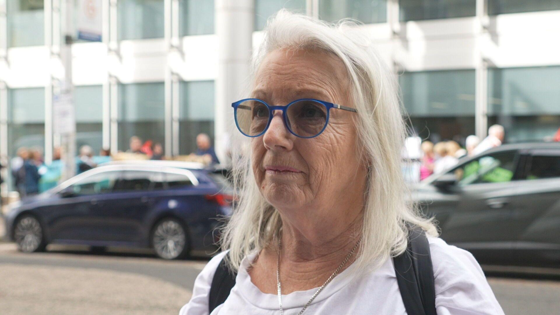 Frances Gilmour, a woman with shoulder length white hair and blue-framed glasses, is standing in a street with -parked cars and pedestrians behind her. She is wearing a white top with black rucksack straps over her shoulders.