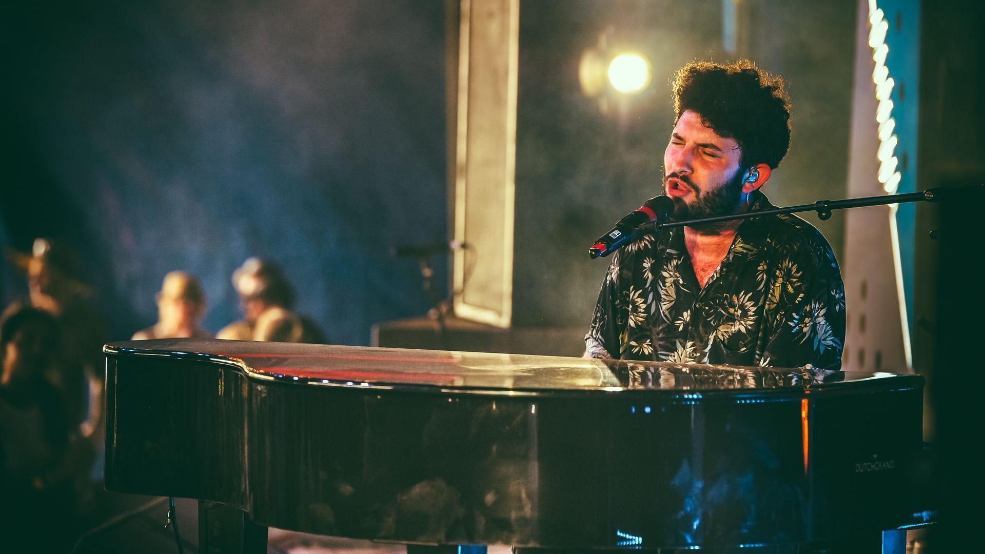 Billy Lockett in a black floral shirt sings at a large black piano