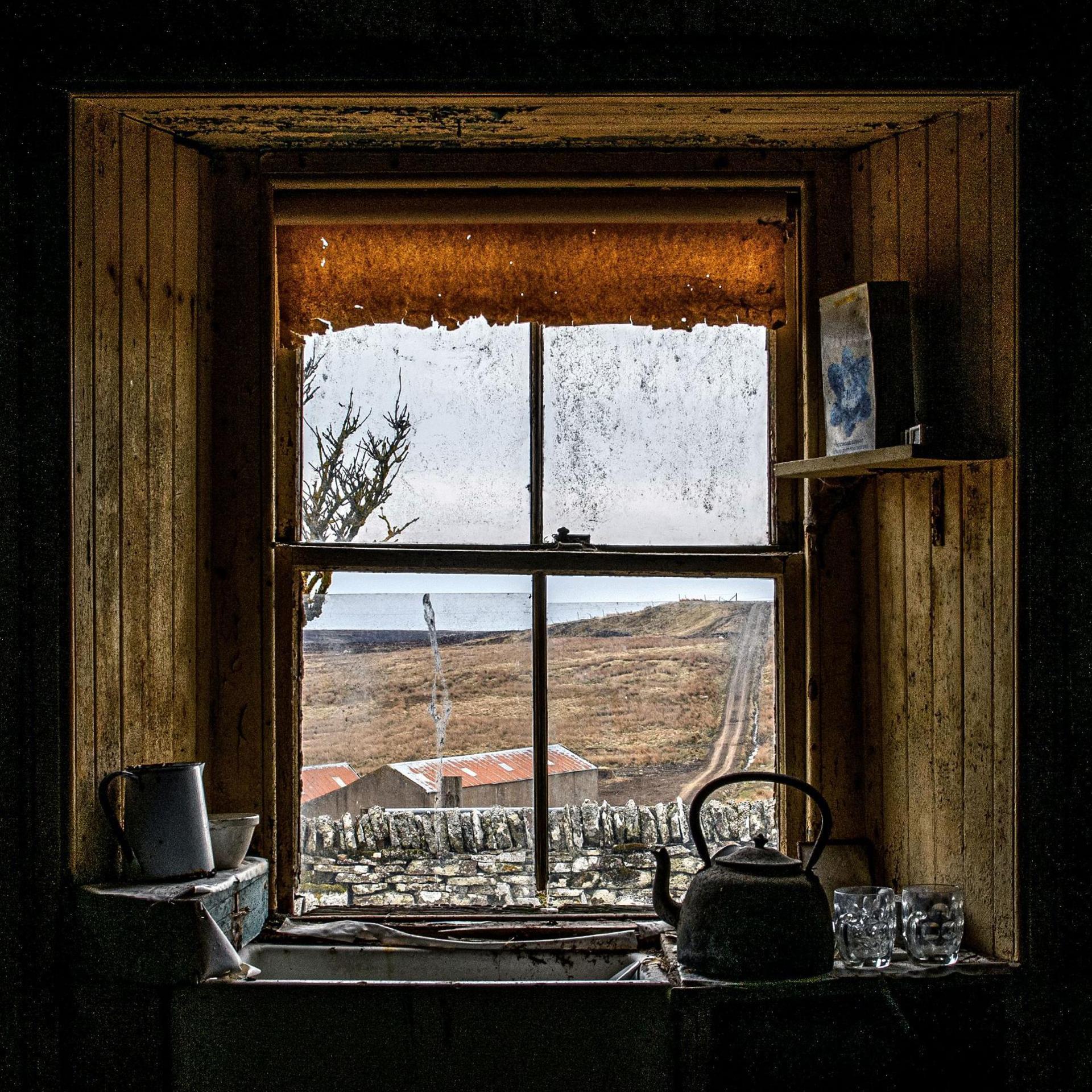 Looking out through a window of the abandoned house. The window has a tattered blind. There is a sink and on it an old kettle.