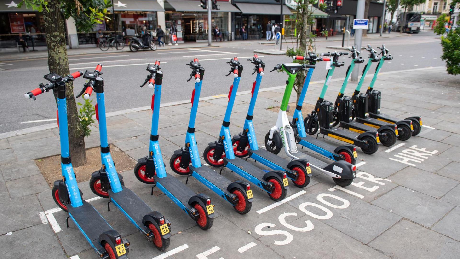 A row of e-scooters, all facing away from the camera, parked in a bay marked out with white lines in a town centre.  The writing on the pavement around the parking bay reads "e-scooter hire"