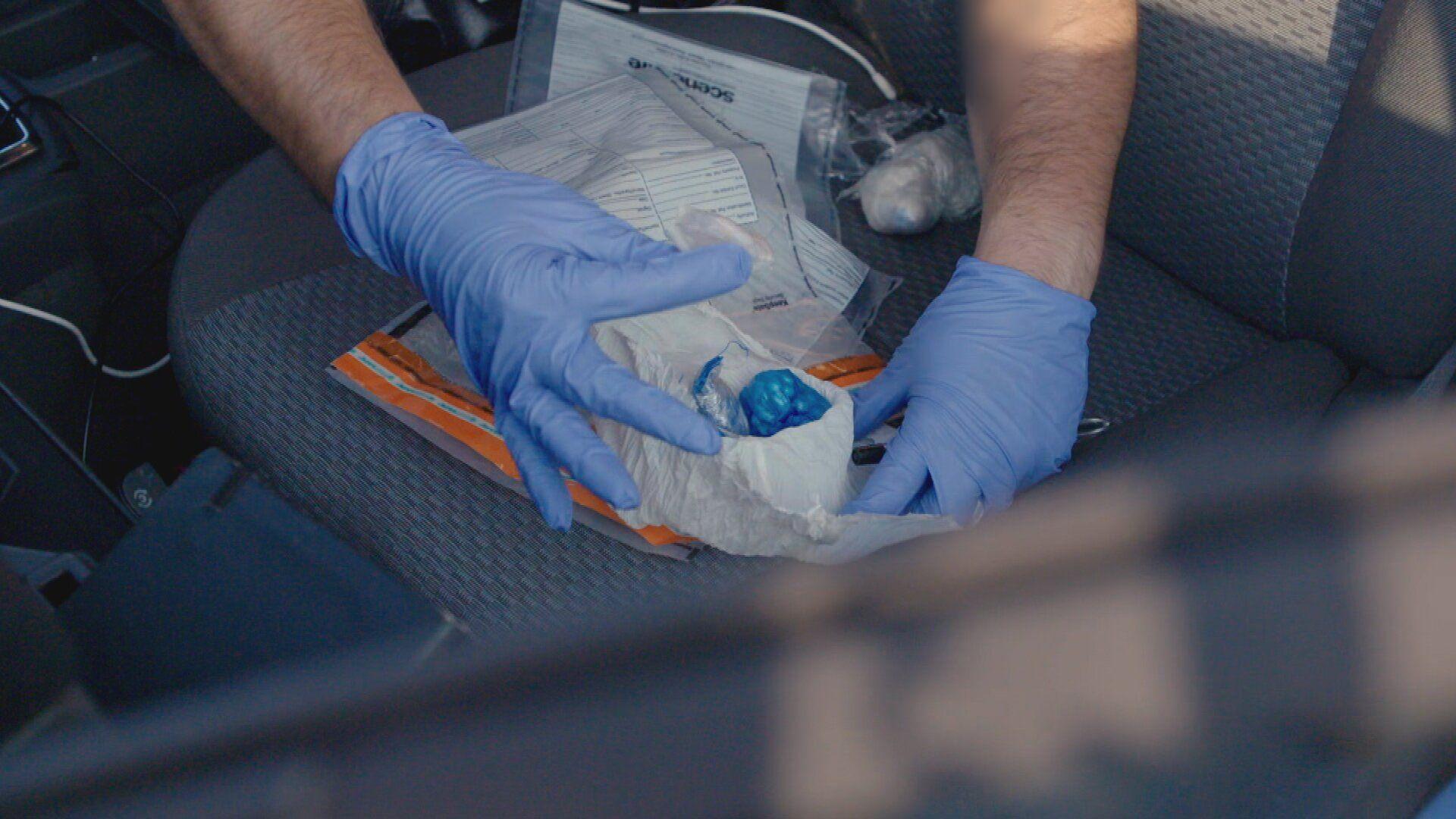 Drugs in various plastic bags and wrappers are being examined by a man wearing blue gloves. It looks like they are on a seat inside a car