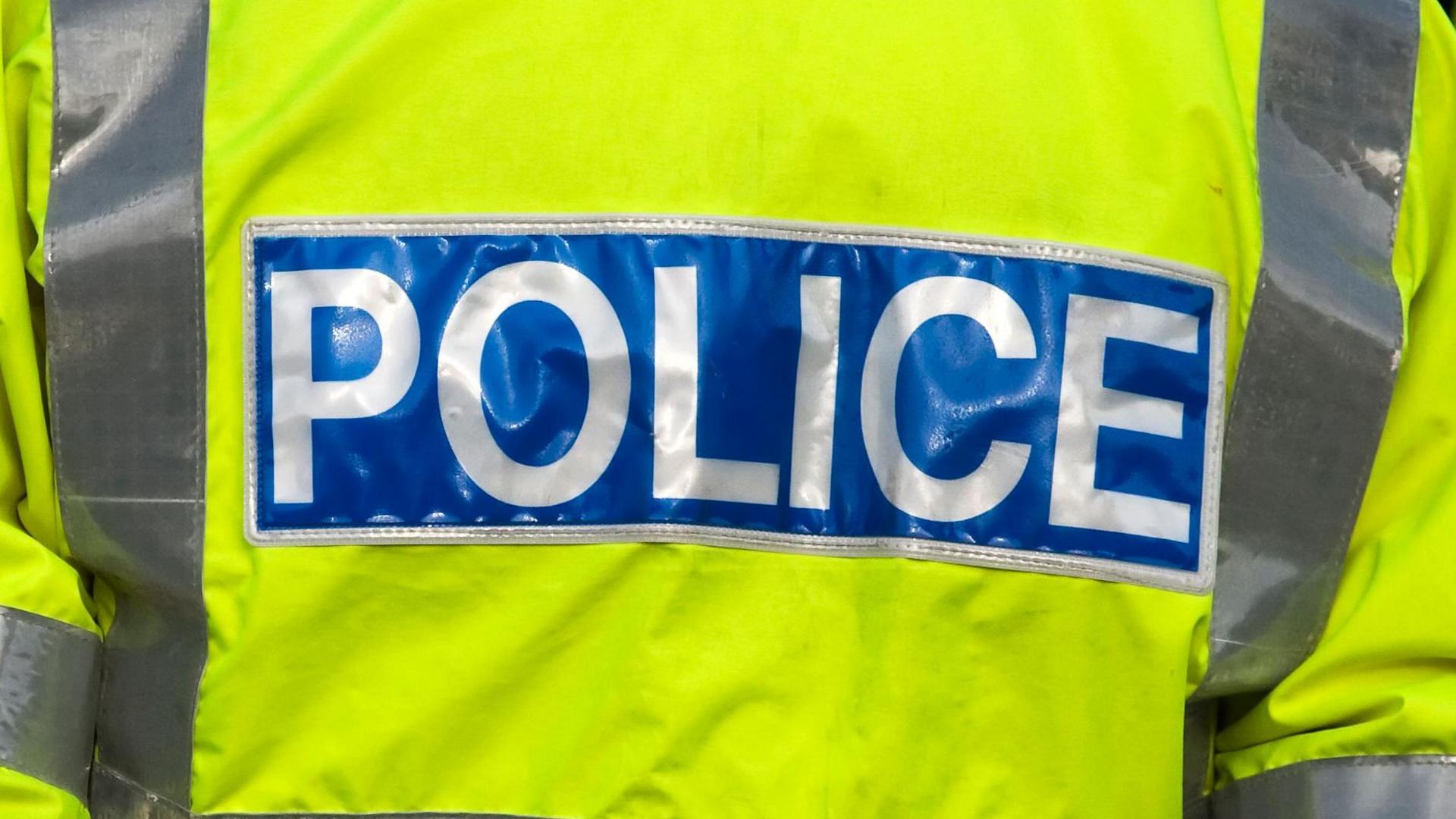 The back of a police officer's yellow high-vis jacket. There is a blue band at the back with "POLICE" written on it in white text.