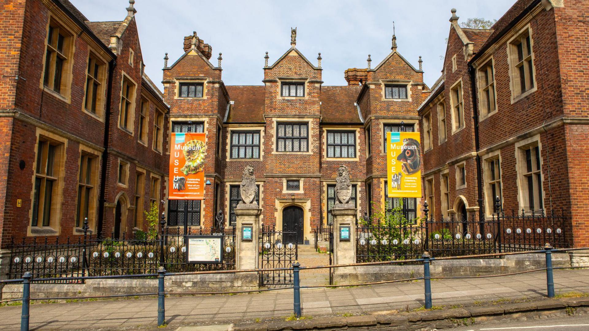 The image shows the outside of a museum, which has two banners on either side of the building: the one on the left is orange with a fossil on it, and the banner on the right is yellow with what appears to be a bird on it. 
There are black fences in front of the main entrance, with a gate in the middle of two pillars. The main black door can be seen in the background down a path.