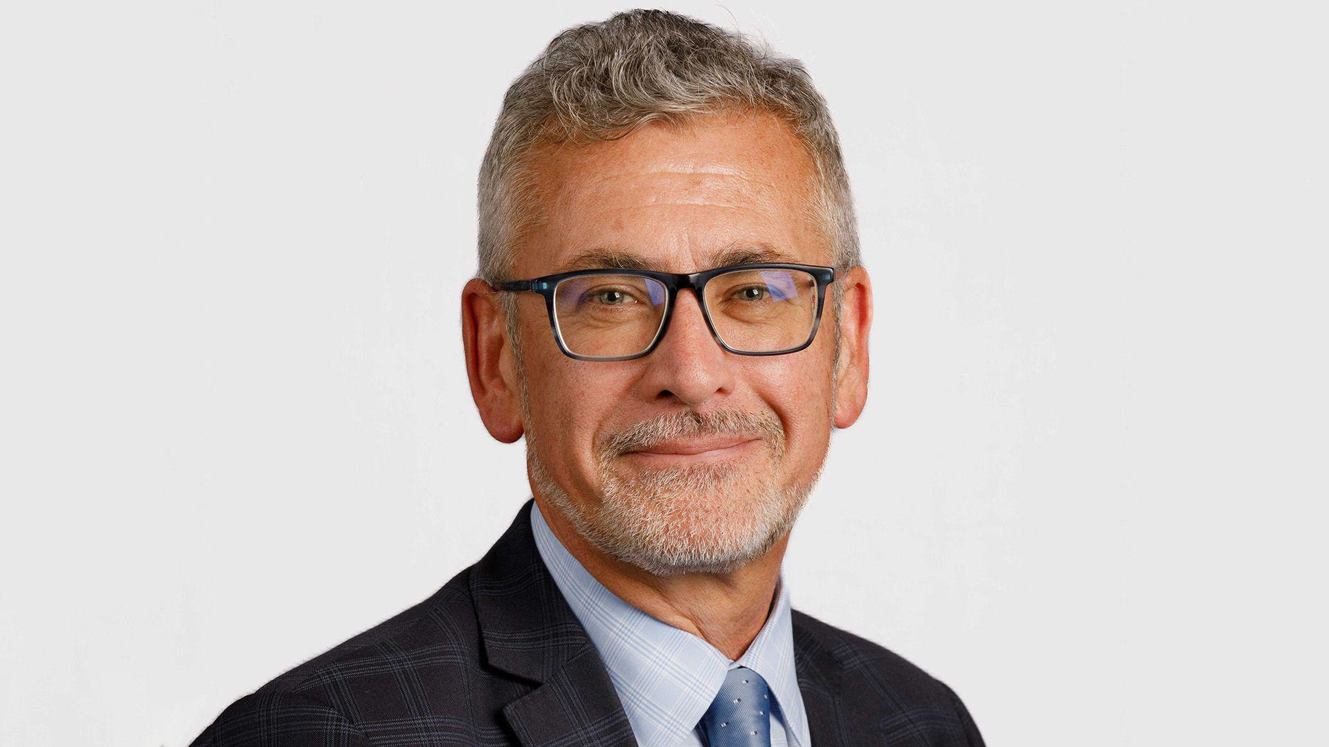 Prof Shane O'Neill is smiling in a university website picture. He has grey hair and beard and is wearing dark glasses, a dark jacket and grey shirt and tie
