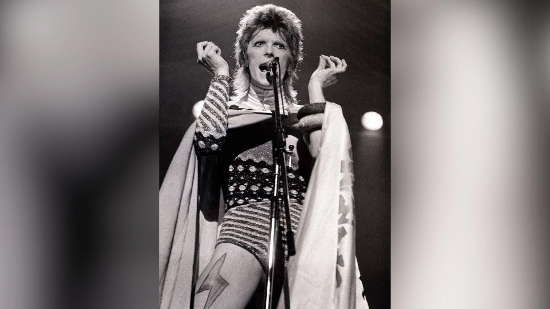 Black and white image of David Bowie performing as Ziggy stardust. He is stood on stage in a bold outfit featuring a lightning bolt on the thigh, in front a microphone.