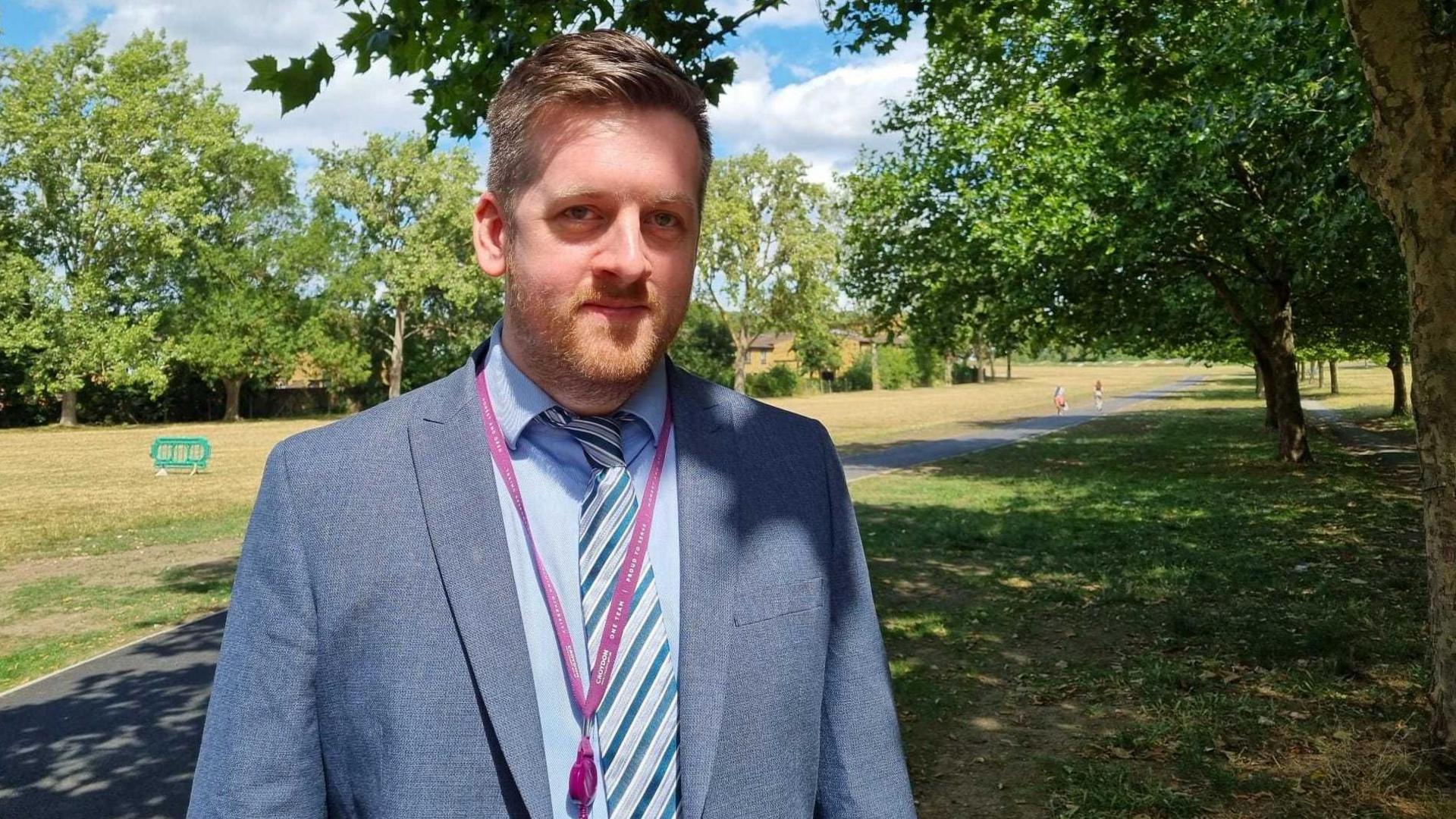 A man with brown hair and a ginger beard is standing in the foreground, looking at the camera. He is visible from the midriff upwards. He is wearing a grey suit with a light blue shirt, a grey-green striped tie, and a light purple lanyard. He is standing in a park, with a narrow grey tarmac path cutting through a field of yellow-green grass. There are trees a short distance from the path on both the left and right. It is a sunny day, with blue skies and a few white clouds in the sky.