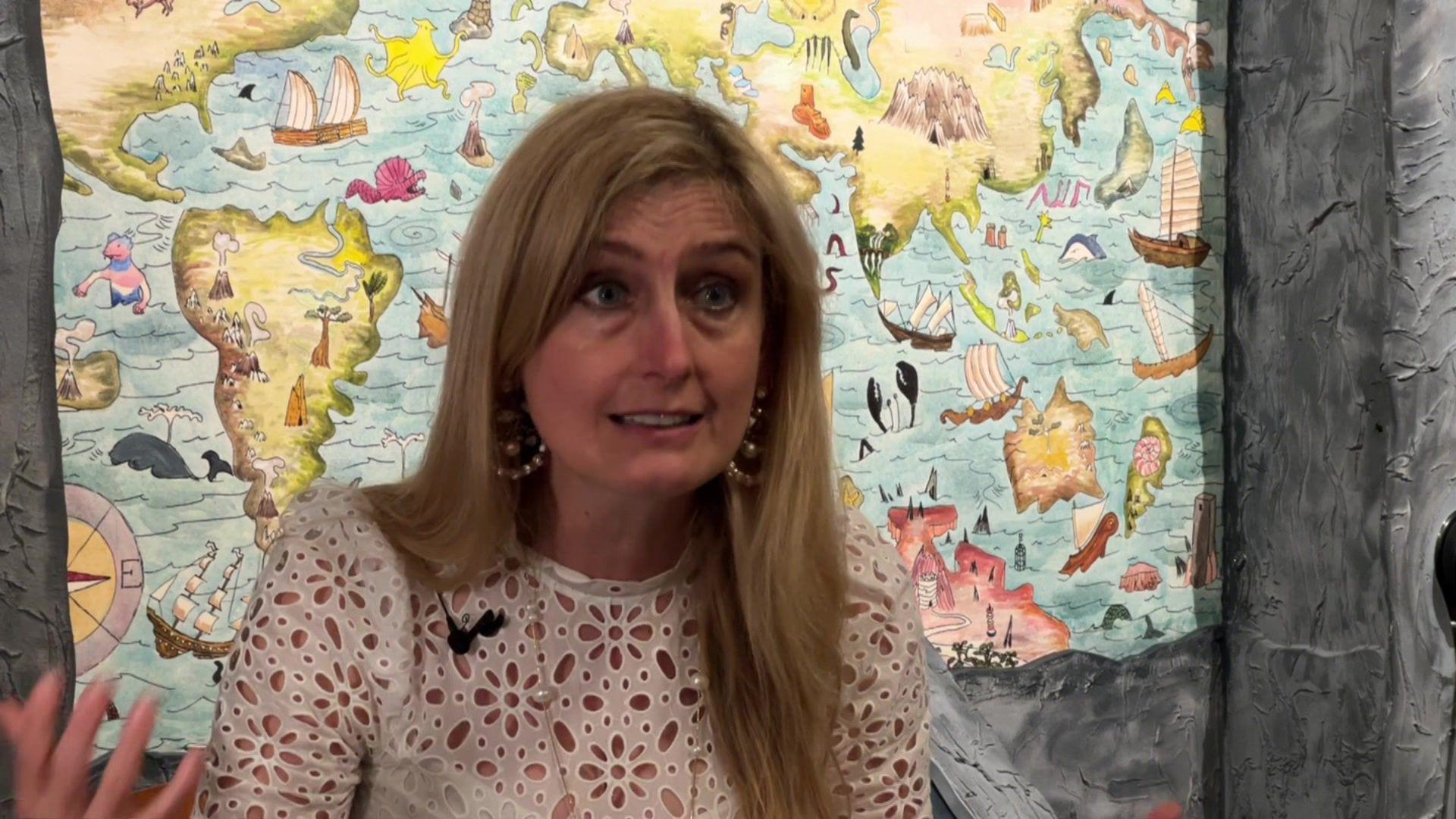 Cressida Cowell giving an interview in front of one of the exhibition's maps. Her hair is down and she wears a white flower top.