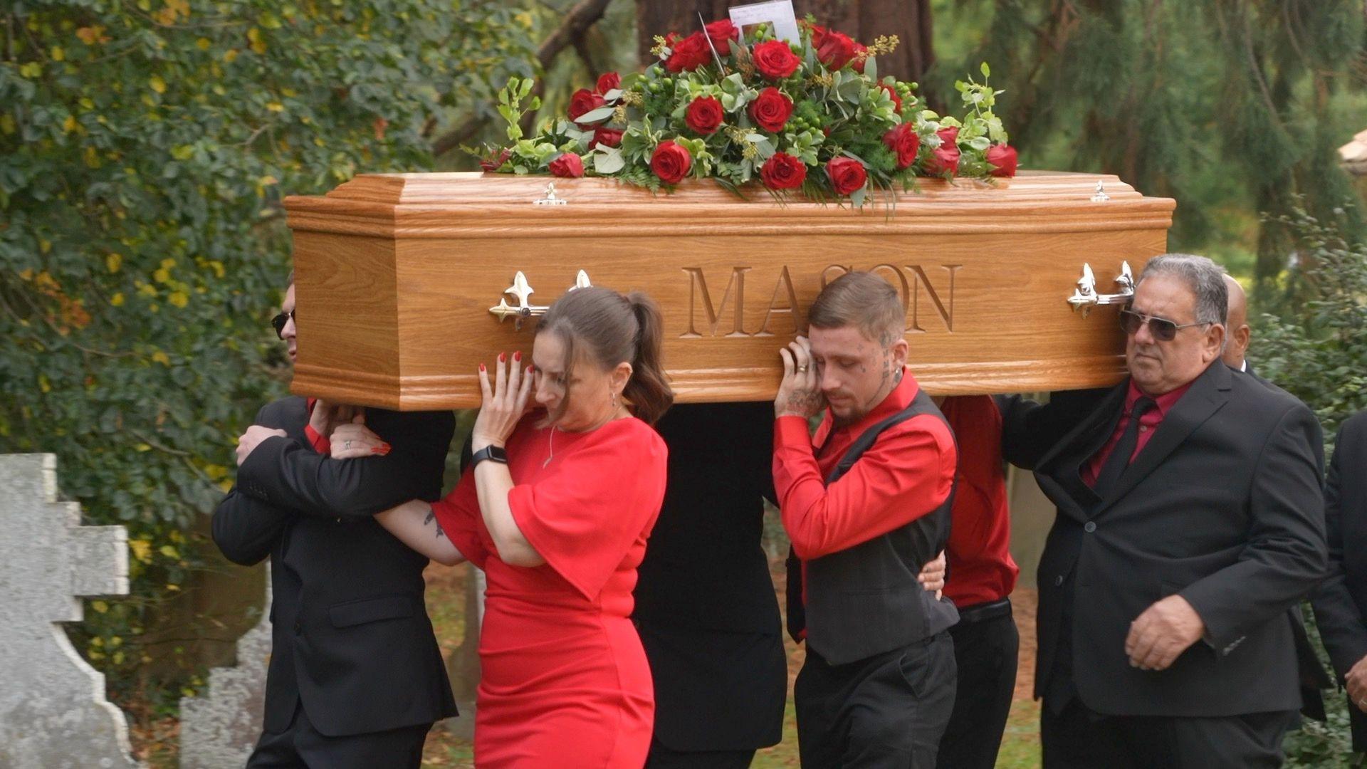 A brown coffin with the name Mason engraved on there being carried by people wearing black and red formal wear.