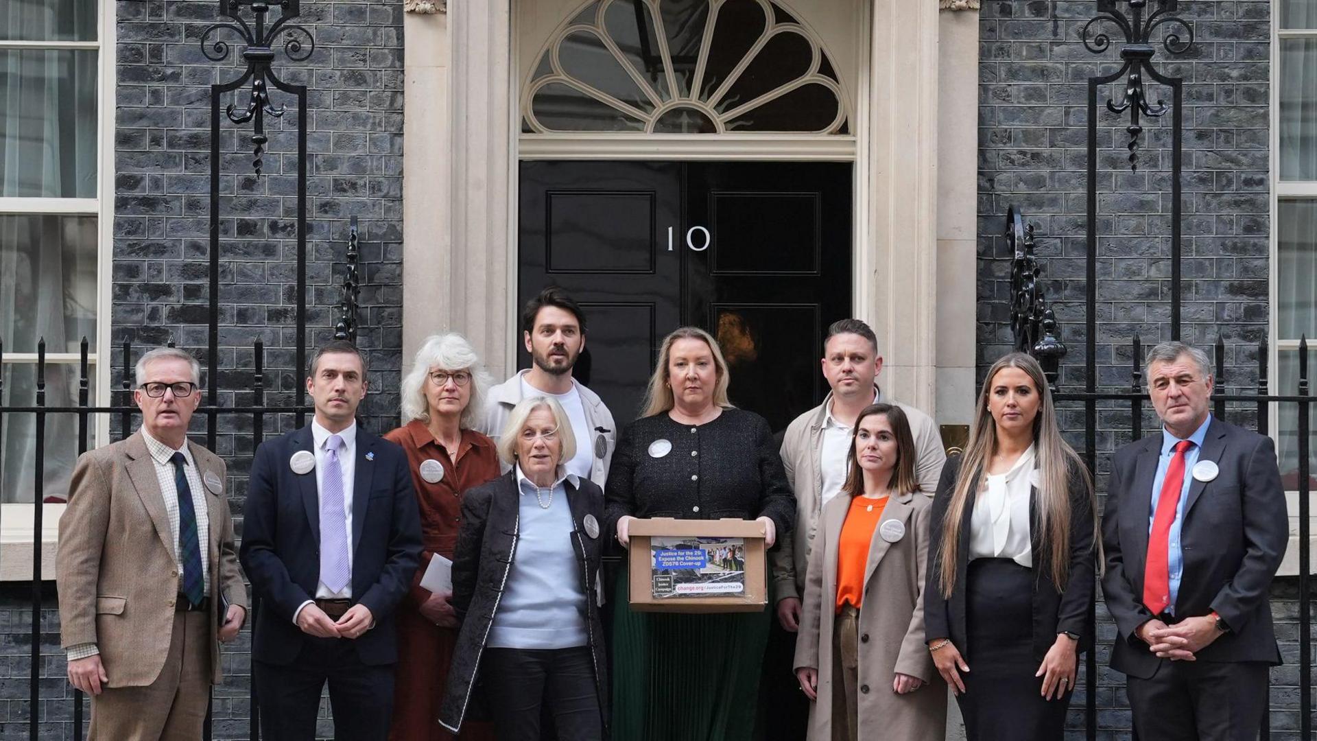 A a group of people are lined in front of Number 10 downing street's famous black door, windows and railings.