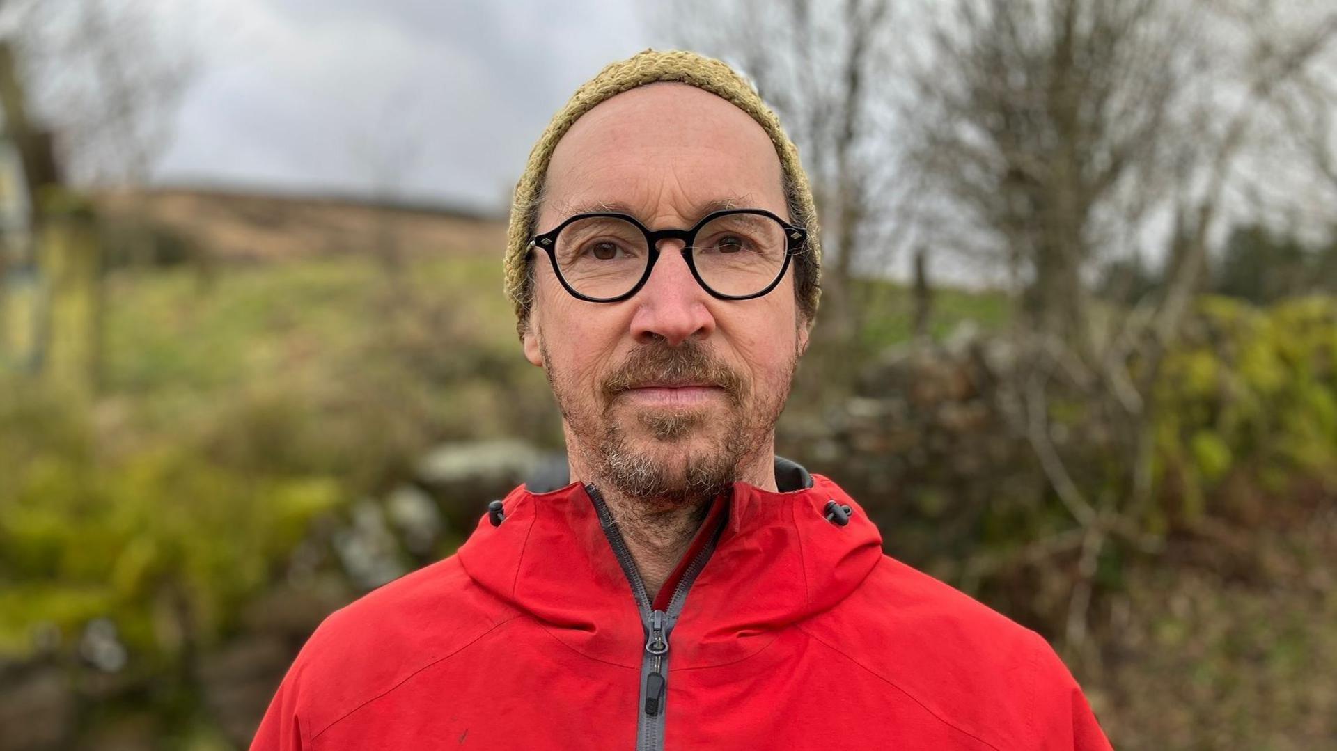 Andy Mather who has brown eyes and beard, wearing a yellow woolly hat, black glasses and red coat outdoors in the countryside