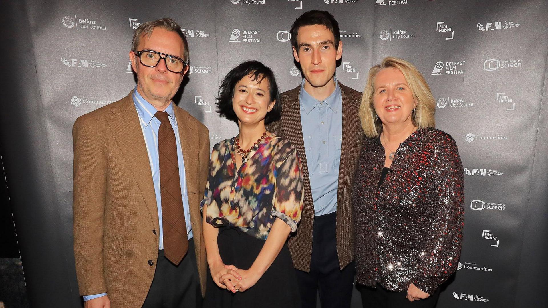 Glenn Leyburn, Lisa Barros D'Sa, Éanna Hardwicke and Michele Devlin standing together looking directly at the camera, all of them are smiling. Glenn has white short hair and is wearing glasses, a light brown suit, blue shirt and brown tie, Lisa has short dark hair and is wearing a red, black, cream patterned top and black skirt, Éanna has short dark hair and is wearing a brown suit and light blue shirt, Michelle has long blonde hair and is wearing a sparkling silver top with black trousers.