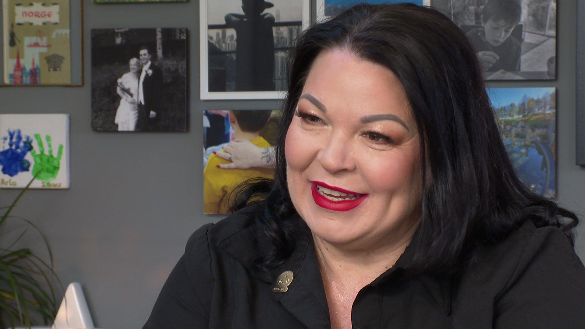 Kiki Deville, with black hair, red lipstick and wearing a black shirt, speaks to the camera