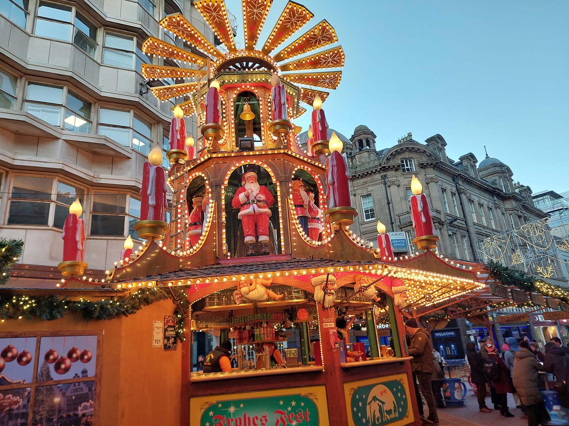 A stall selling alcohol at a Christmas market. The wooden stall is adorned with golden fairylights, giant fake red candles, a bell, Santa decorations