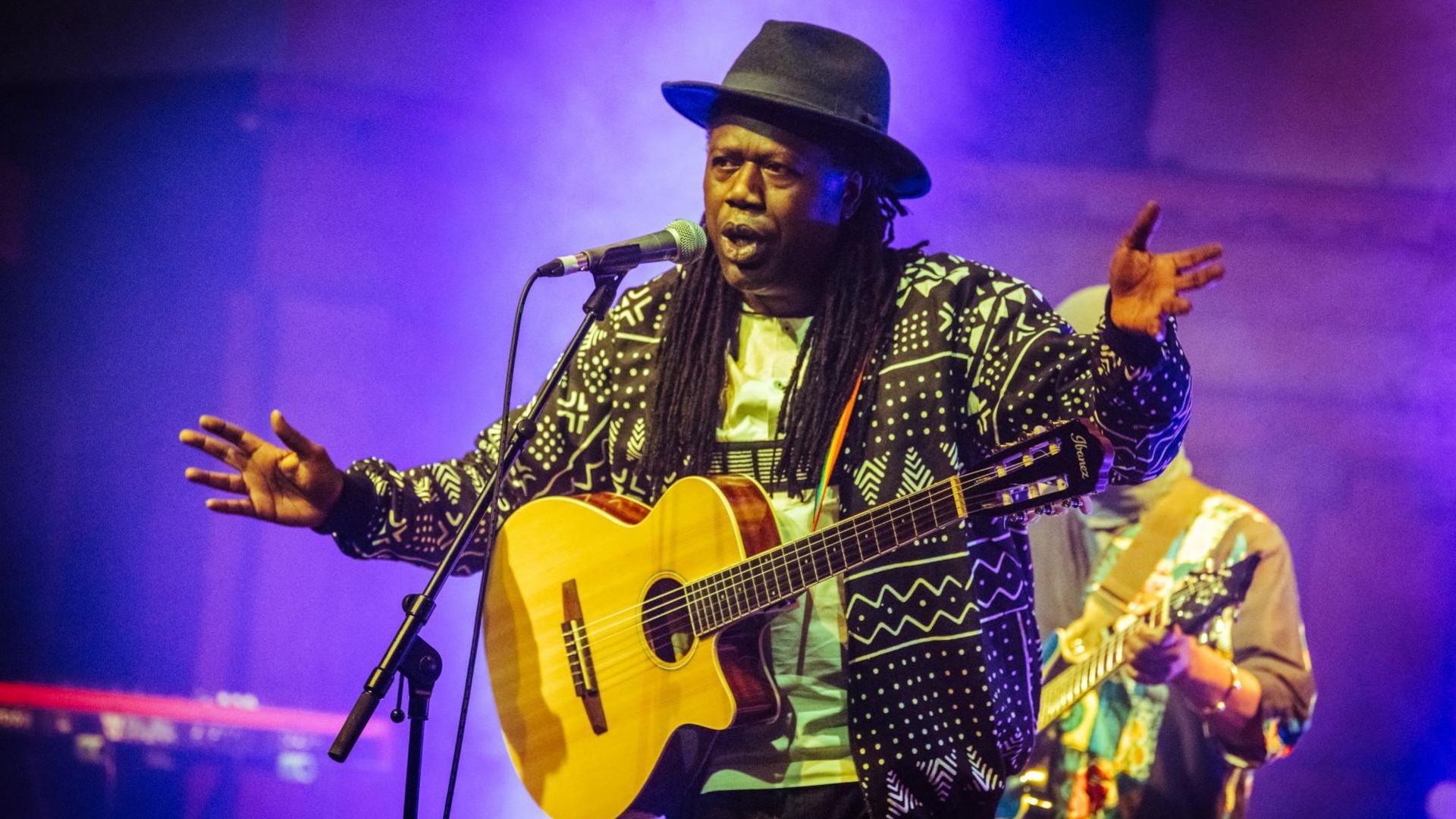 A band member, who is wearing a black fedora, and with a guitar slung across his shoulders sings into a microphone. He is wearing a brightly coloured coat.