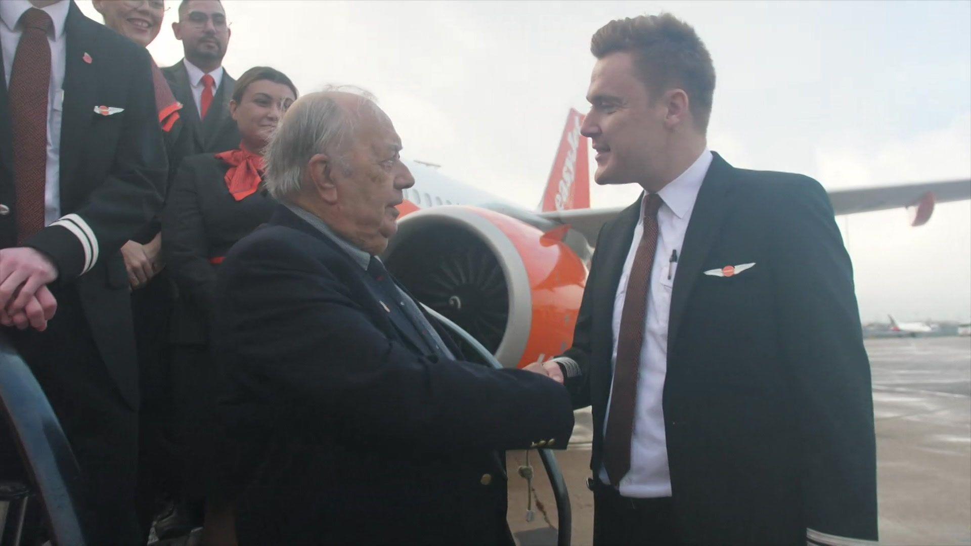 An older man is shaking hand with a younger man on the steps of a plane. Both are smartly dressed.