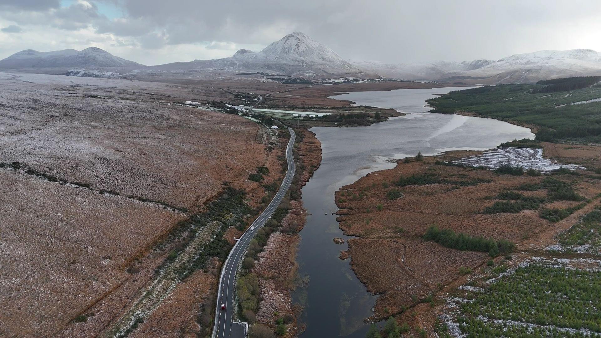 An aerial shot of County Donegal. There is a long stretch of road on the left of a large river which stretches the length of the road. There are large fields on either side. In the distance is a snow-dusted Mount Errigal.