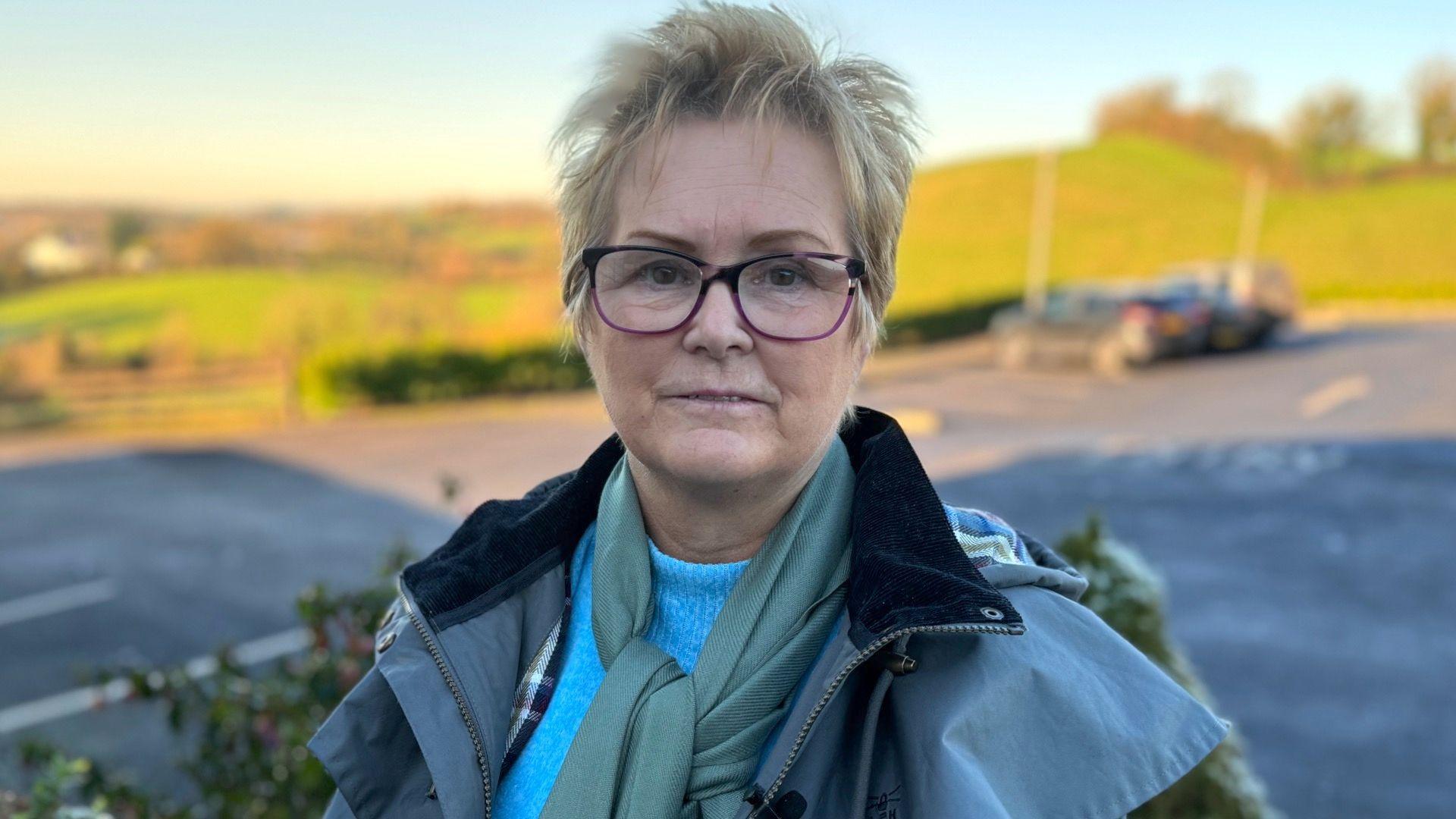 Christine McFlynn has short blonde hair and glasses. She is wearing a blue jumper, green scarf and a Barbour-type coat. She is standing in what looks like a car park with green field behind her.