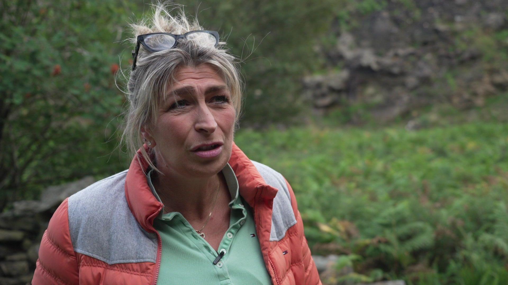 A woman with medium length greying hair looks off camera. She wears an orange and grey jacket and a green top. She has glasses perched on the top of her head. She stands with vegetation and a stone wall in the background.