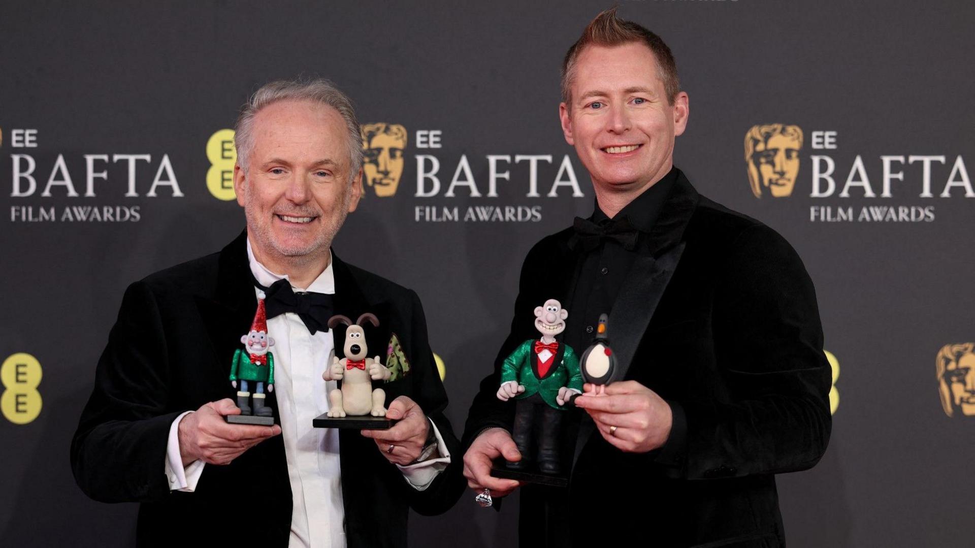 Two men wearing suits and bow ties hold small Wallace and Gromit figurines. They are standing on the Baftas red carpet.