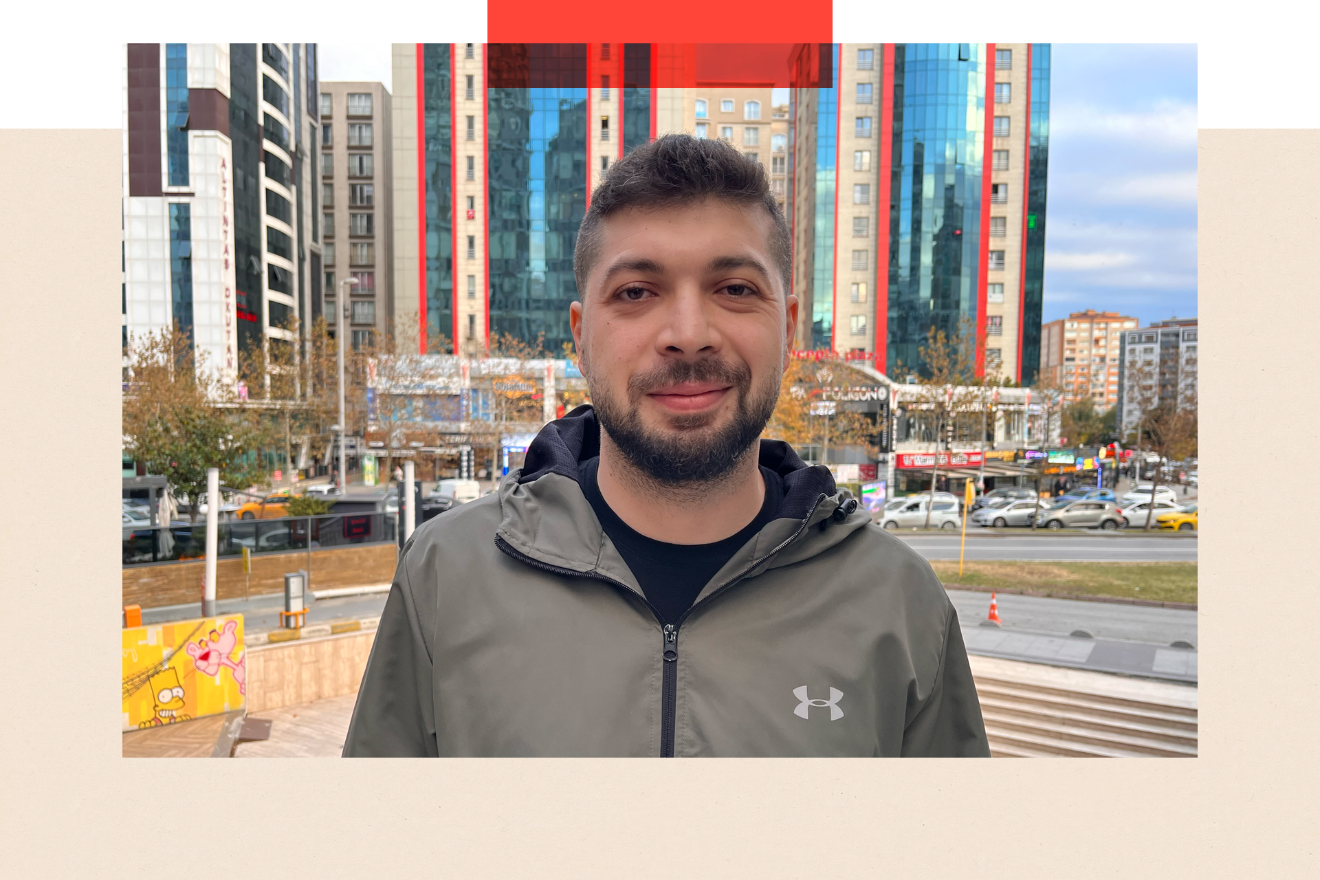 Photo of Hussam - a young adult man with a moustache and trimmed beard. He is staring straight to camera. Behind him is a road and some high rise buildings.