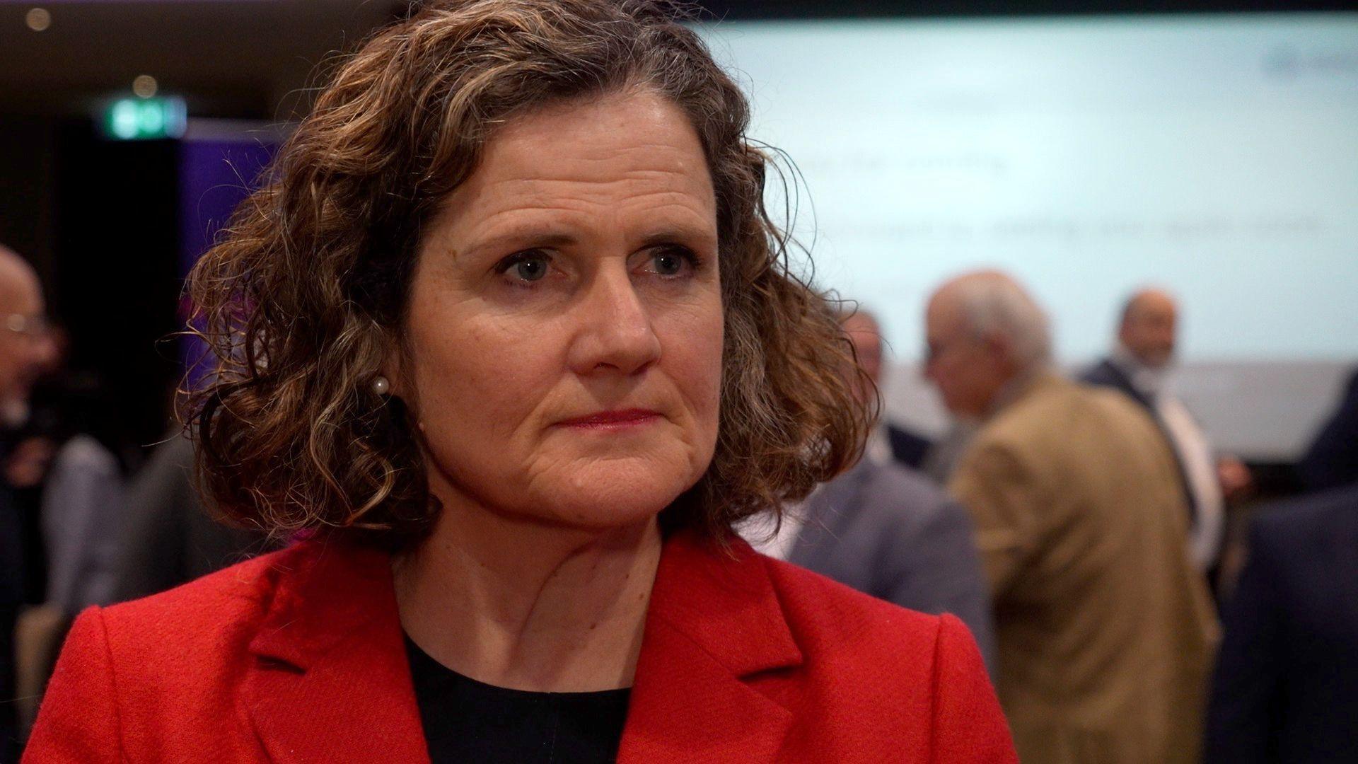 Becky Sherrington is wearing a red blazer and a black top. She has curly brown hair. There are a number of people in the room behind her.