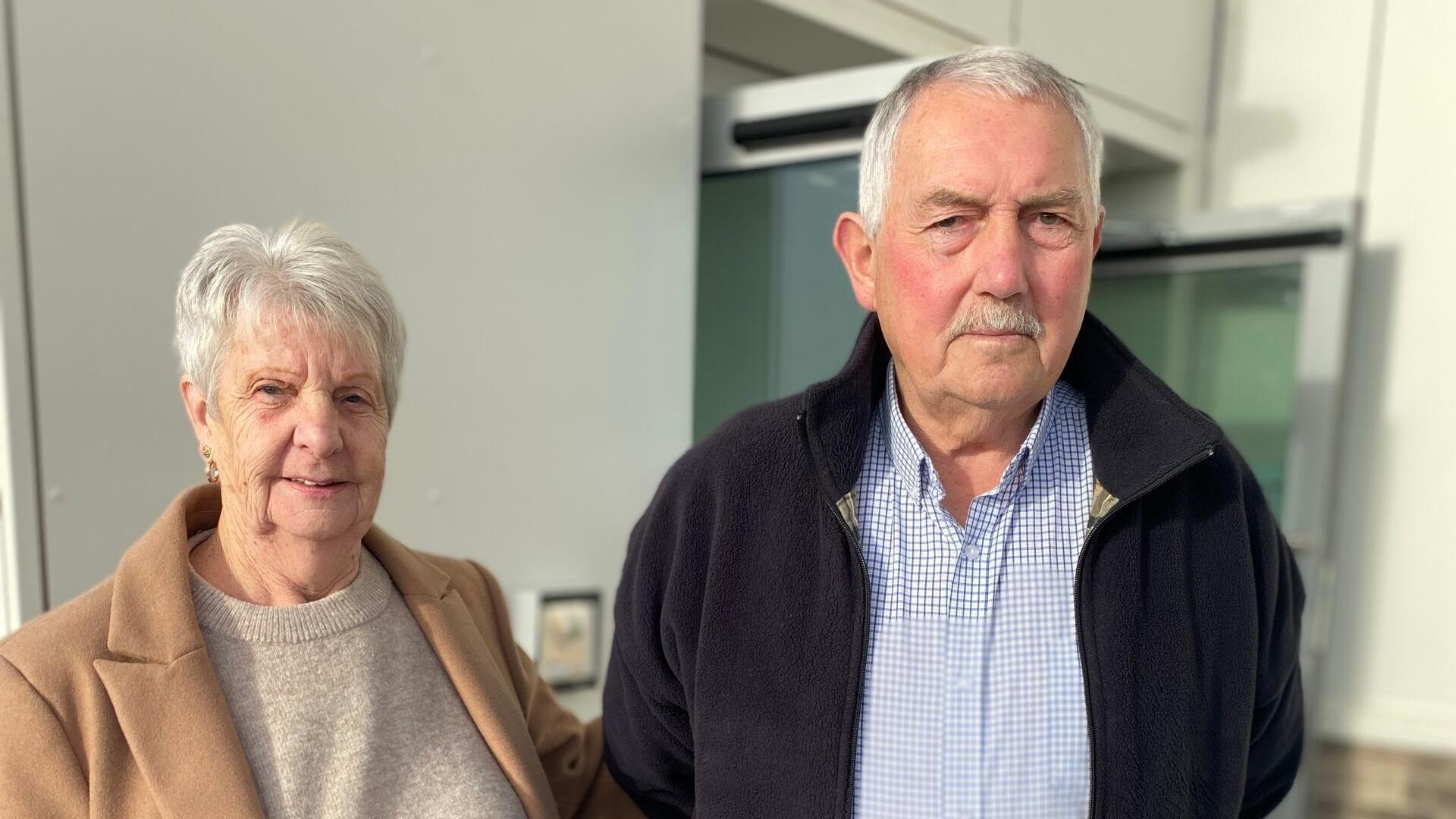 A man and a woman stand outside a new NHS testing centre. The woman has short white hair and wears a light-brown jacket and cream pullover, while the man has short white hair, a white moustache and wears a blue checked shirt and a black fleece.