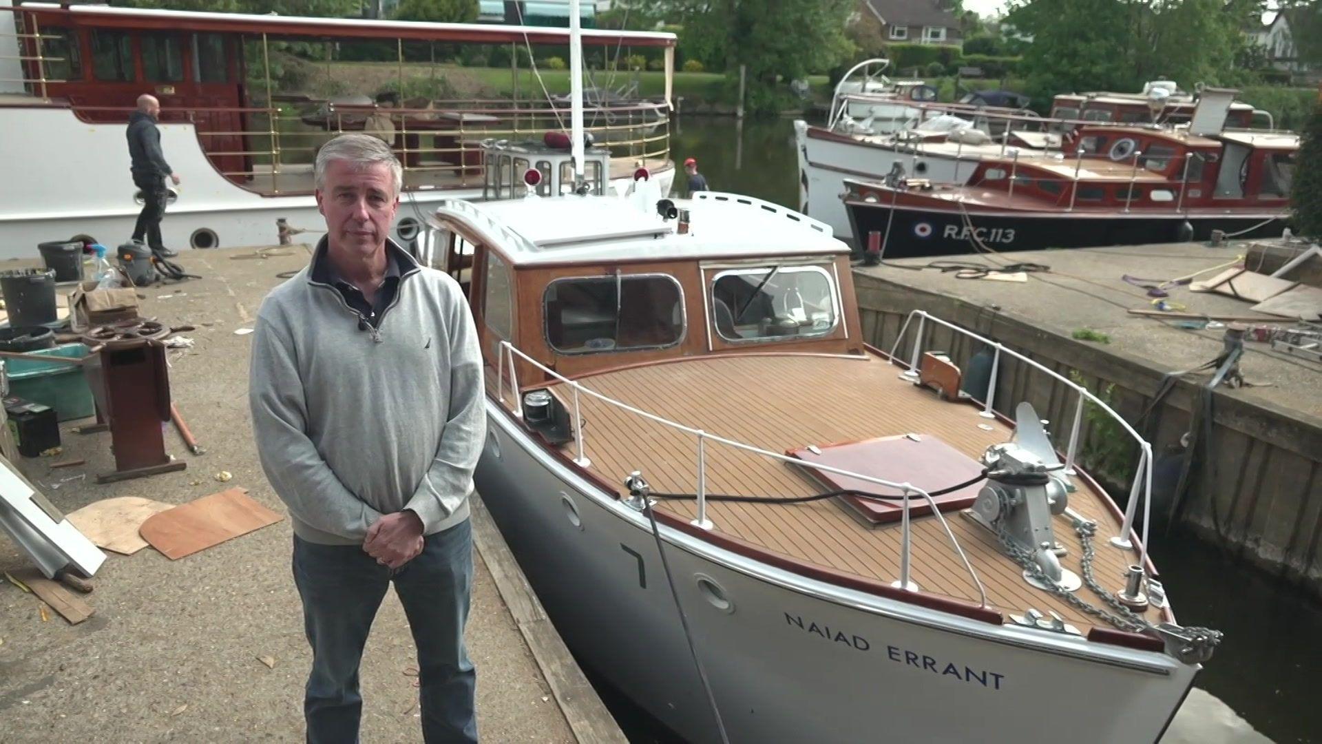 A man with grey hair and a grey jumper stands next to a white ship.