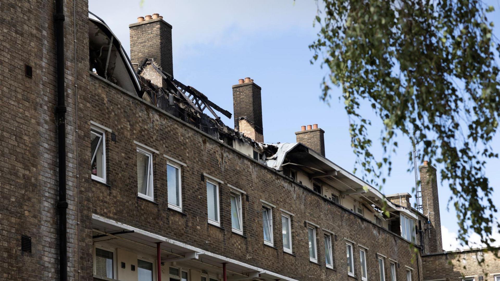 A brown brick residential building, with a roof gutted by flames 