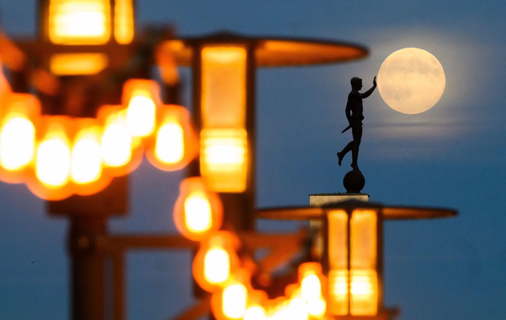 Bright orange lights are seen in the foreground next to a statue of someone with their hand out, which the photographer has lined up to make it look like the statue is touching the Moon, at dusk in Hannover on Wednesday.