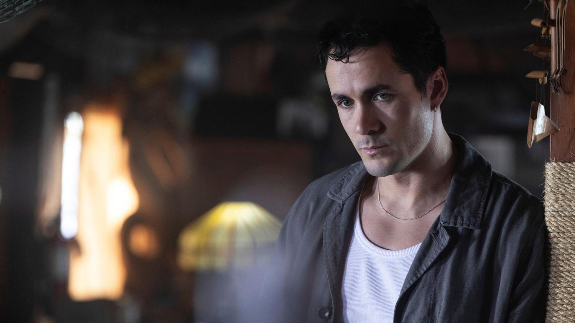 Actor Jonah Hauer-King, in character, leans against a pillar inside a dimly lit restaurant with wood panelled walls. the only illumination is a lamp and shaft of sunlight behind him. He looks troubled as he stares into the middle distance.