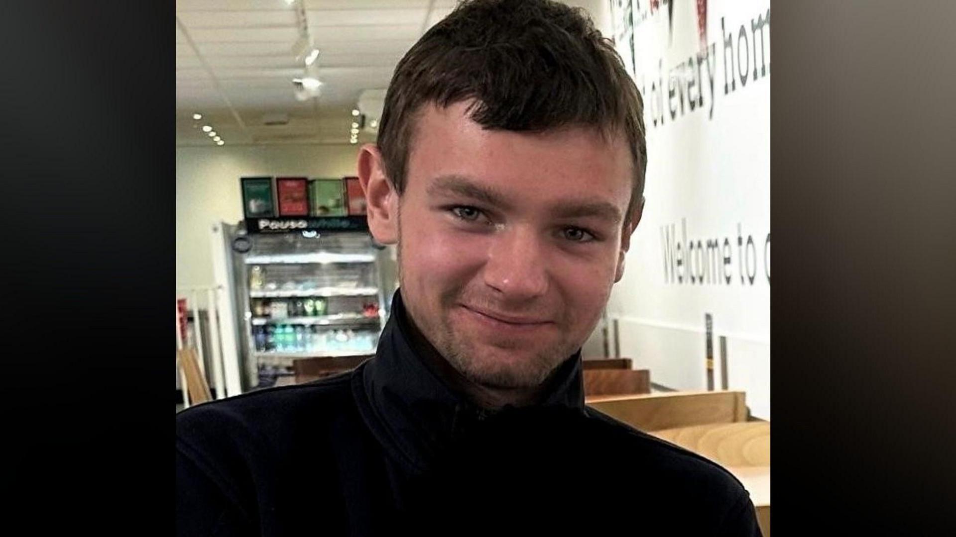 Close-up image of Ben Procter smiling directly at the camera. He is wearing a black jacket and standing in a cafe-like room behind him.
