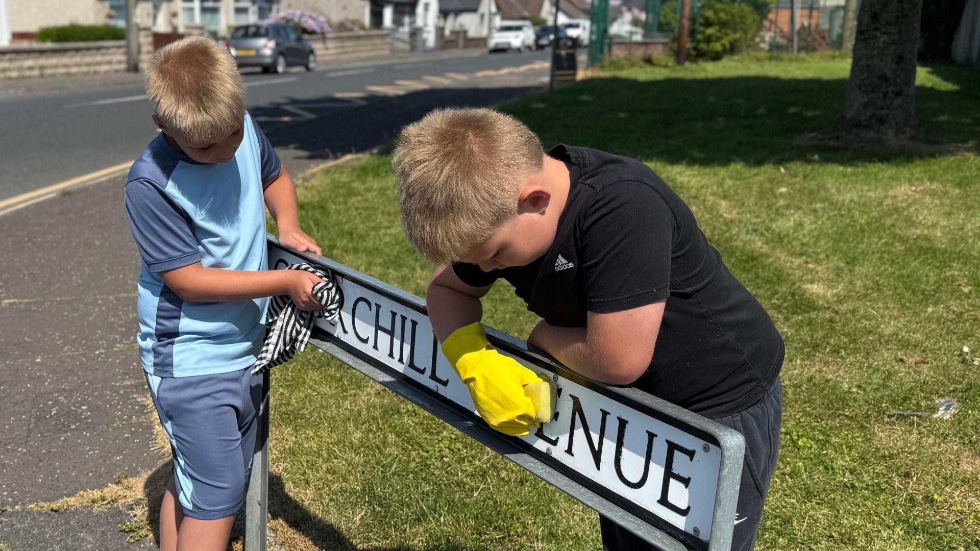 Two children are cleaning a street sign that reads "Churchill Avenue." One child, wearing a light blue shirt, wipes the sign with a checkered cloth, while the other, dressed in a black shirt and wearing yellow rubber gloves, scrubs the sign with a sponge. They are standing beside a grassy verge next to a road. 