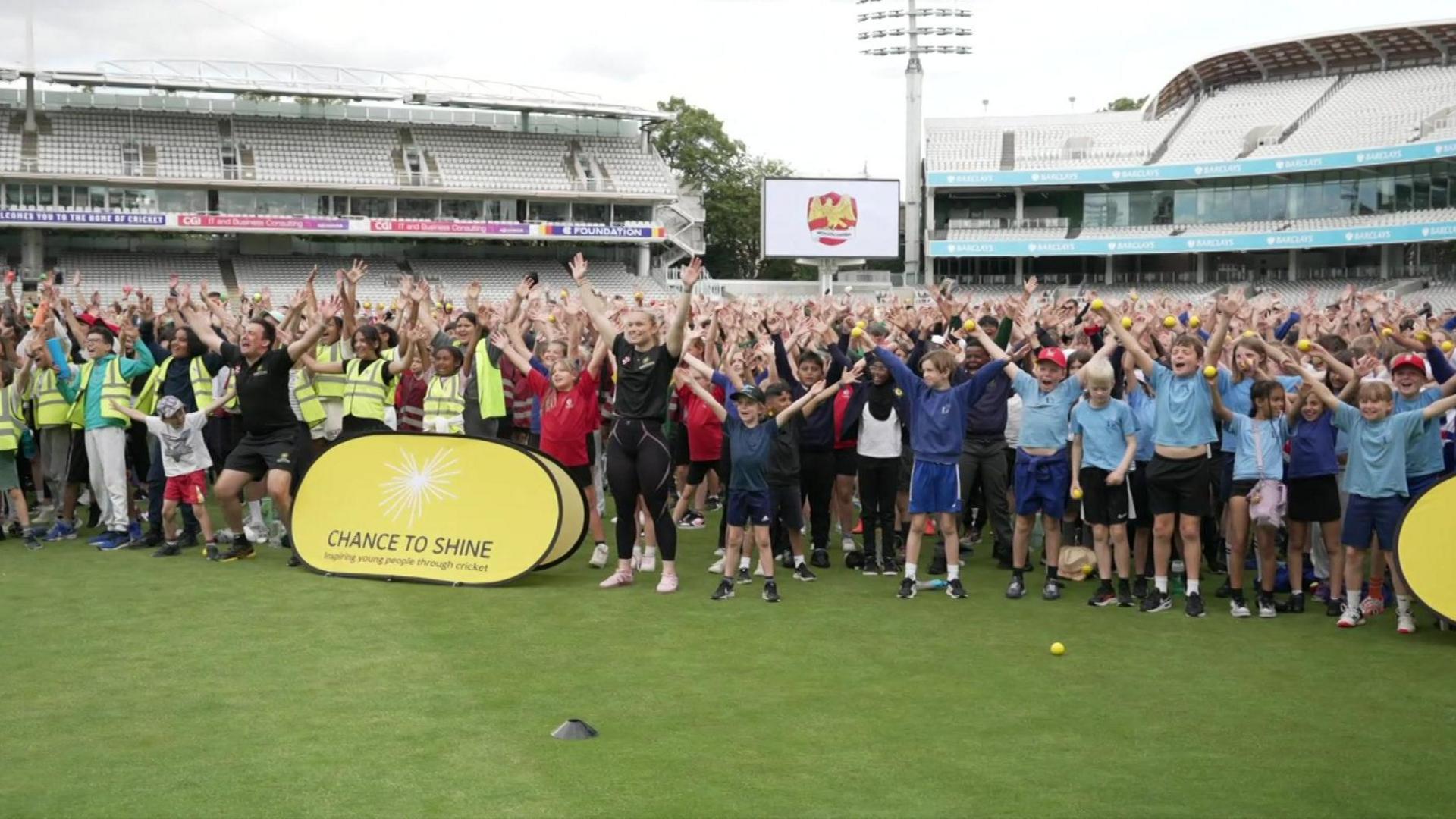 Hundreds of children standing on the pitch at Lord's in a group celebrating, many with their arms in the air. TV Gladiator Fury is in the front row.