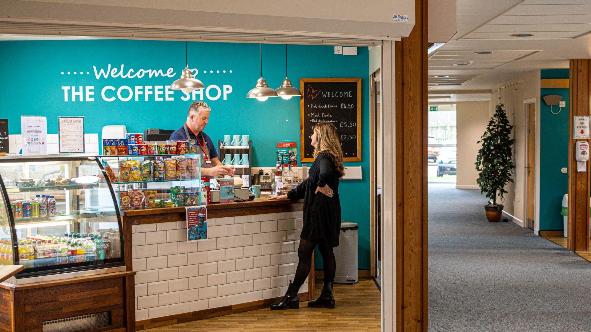 A coffee shop inside Myton Hospice in Coventry. A man is serving a woman. There are a selection of snacks and drinks in a display cabinet nearby. 