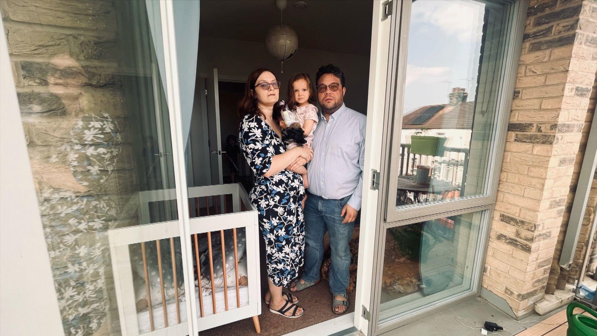 A couple holding a young child stands in their flat’s doorway beside a glass-panelled balcony.