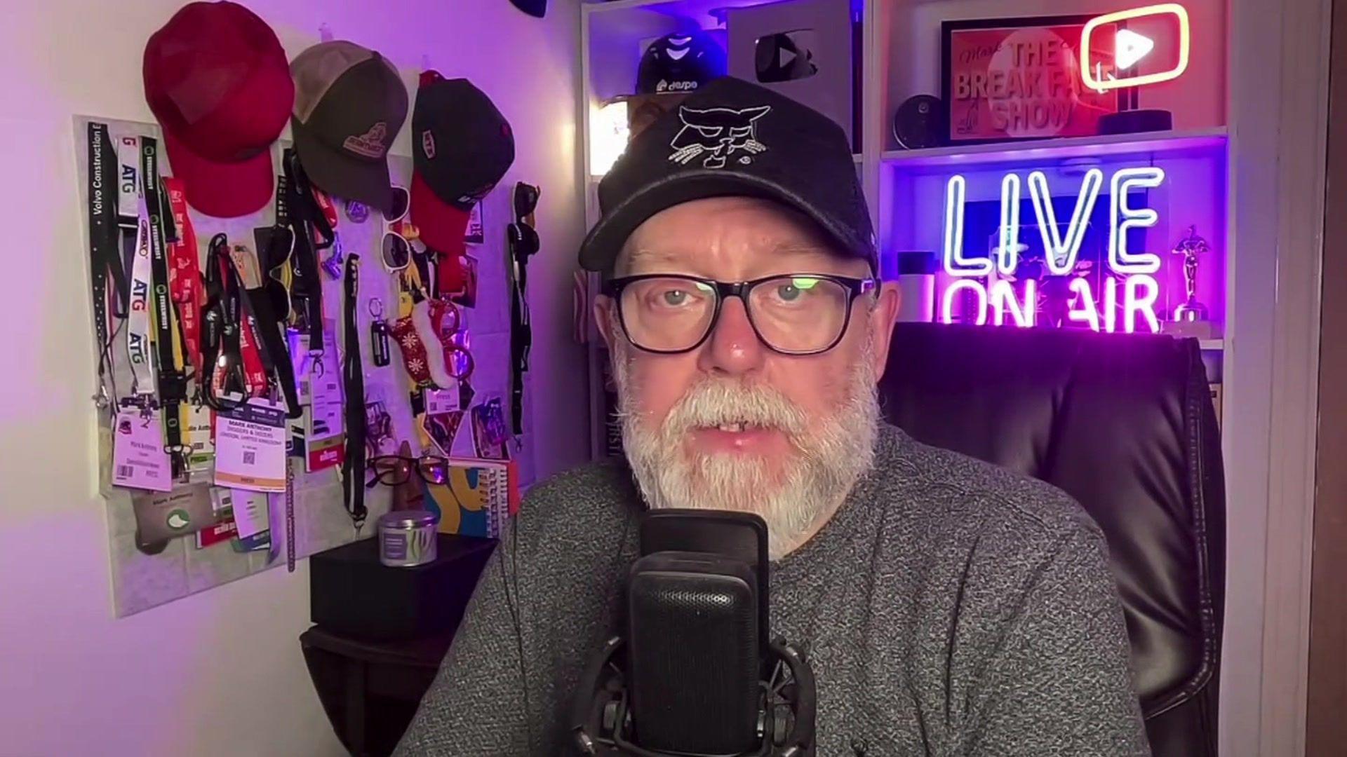 Demolition News editor Mark Anthony in his home studio, talking in front of a microphone. There are hats and lanyards hung on a wall beside him. Behind him, there are illuminated YouTube long sign and LIVE ON AIR sign. He is looking at the camera.