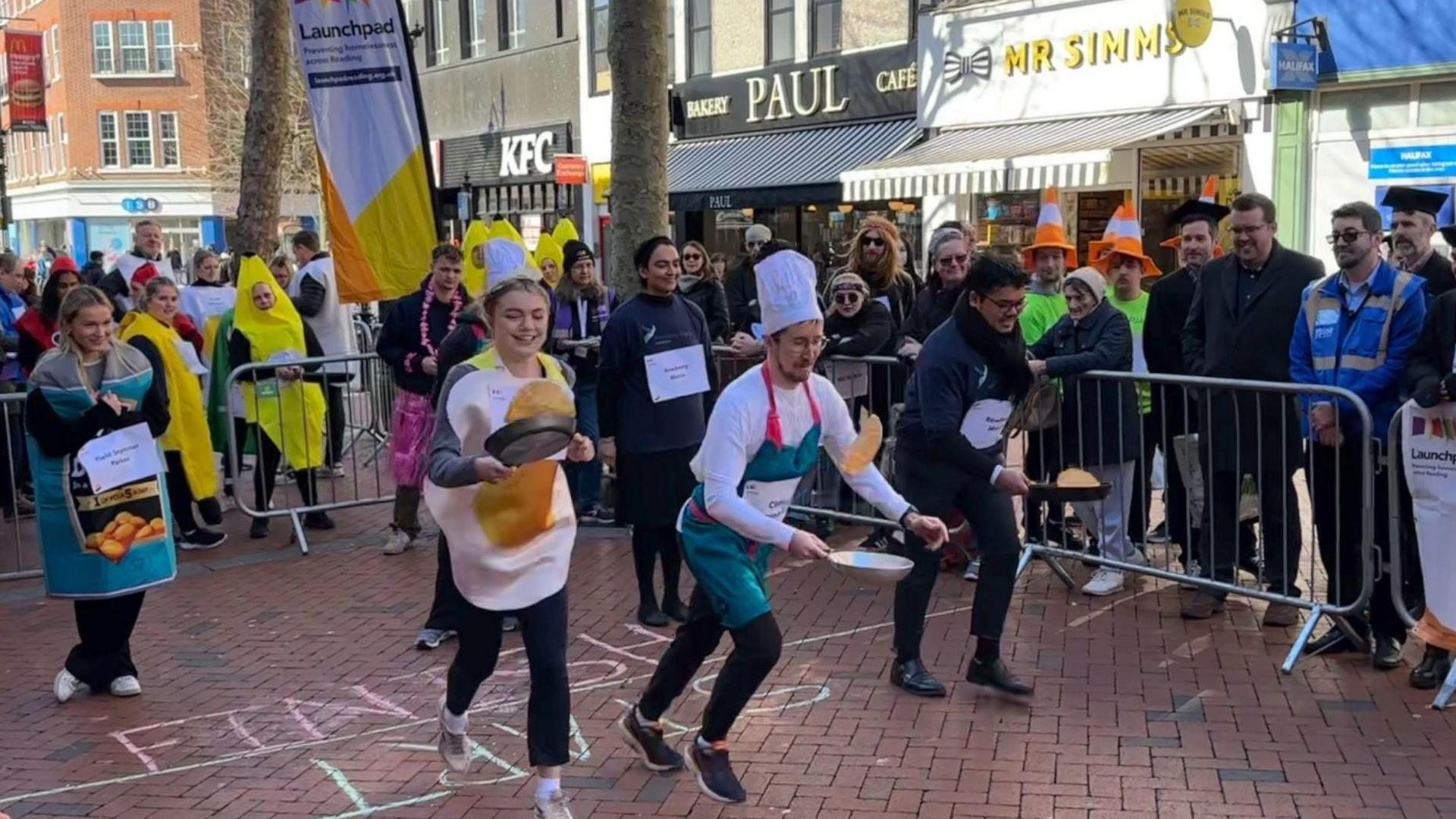 Crowds of people are gathered around watching people set off for a pancake race. The competitors are wearing fancy dress costumes, with one man dressed like a chef with a tall chef's hat. A young woman is dressed as a fried egg and another in a baked bean tin costume. They are holding frying pans and attempting to flip their pancakes as they run.