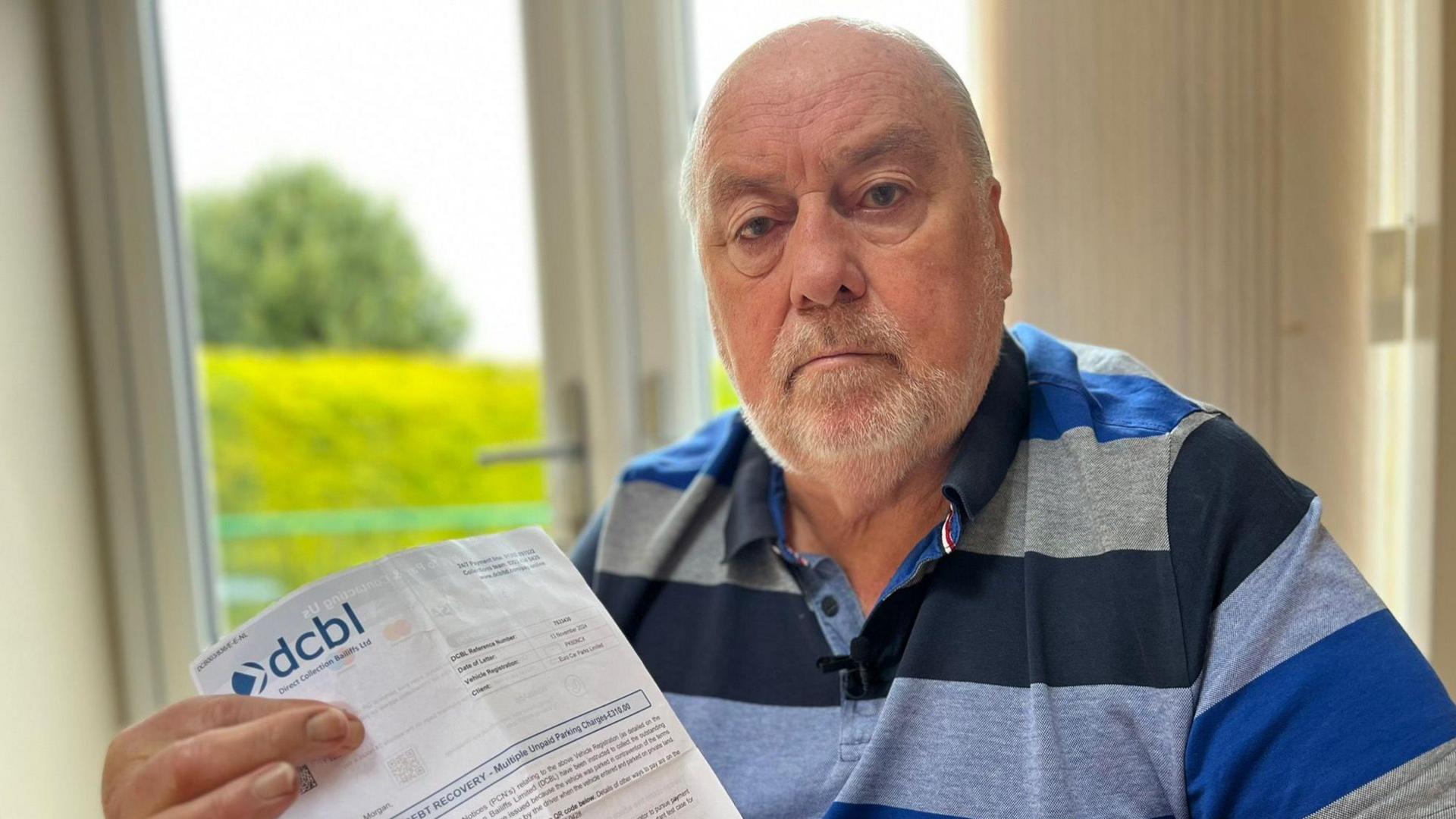 A bald man with a stubble beard, wearing a blue striped polo top and holding a letter from Direct Collection Baliffs Ltd