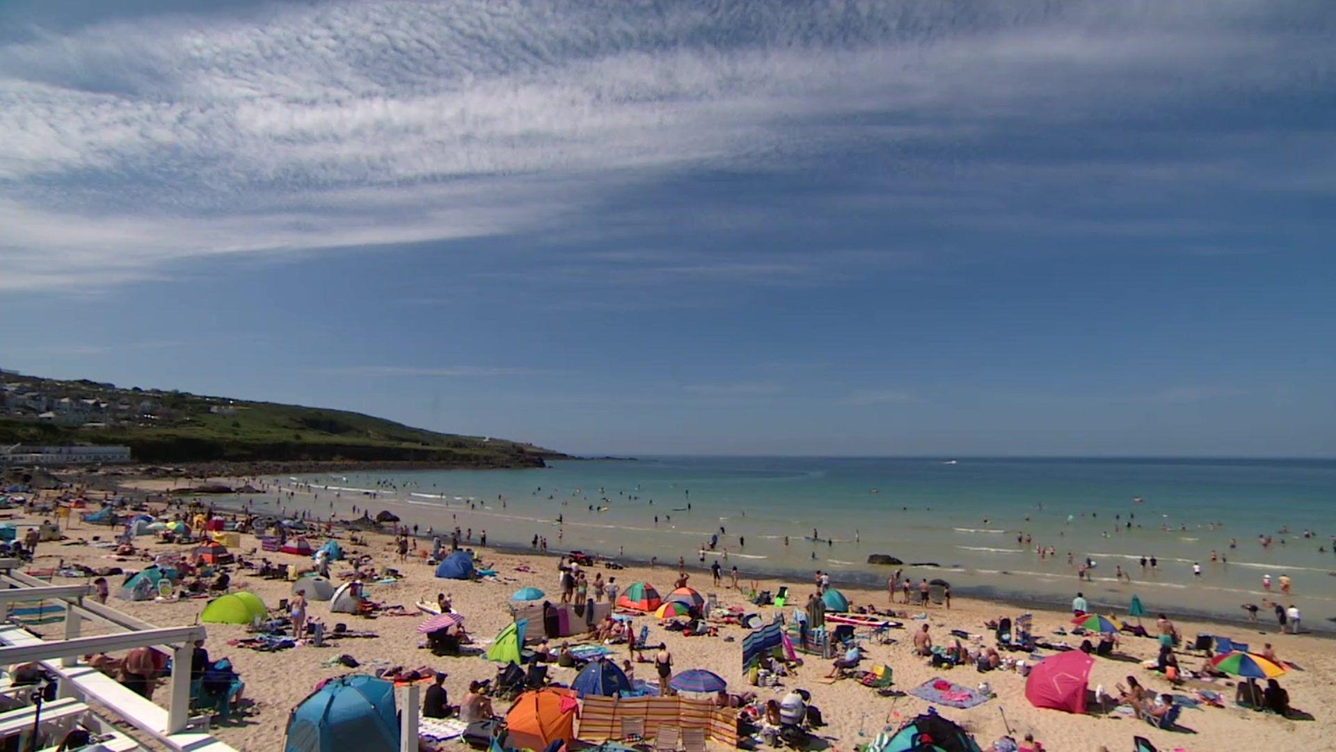 A photo of Porthmeor beach. It shows hundreds of people on the beach, with some people swimming and paddling in the sea. There are towels, tents and umbrellas on the beach, with people sunbathing. The sky is blue.