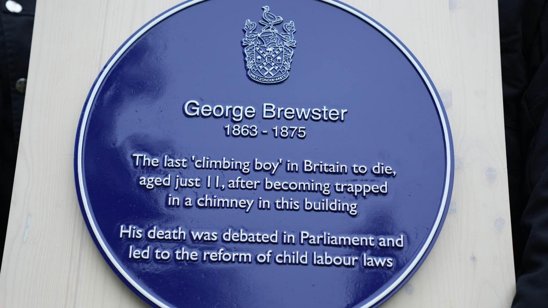 A blue plaque is seen on a white background. In white writing it commemorates George Brewster, born in 1863 and who died in 1875. It makes reference to his death and a subsequent debate in parliament that led to child labour reforms