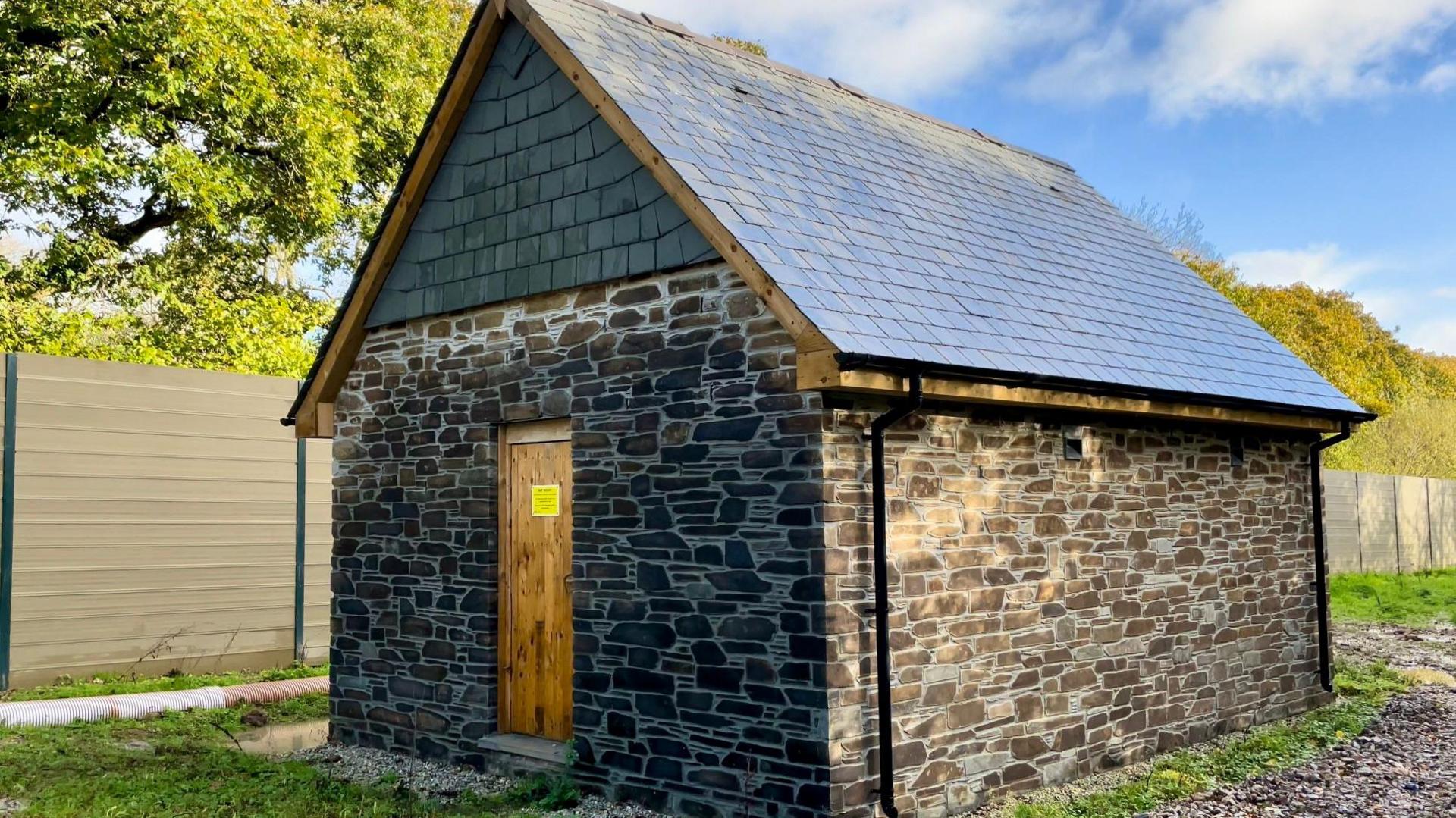 A stone barn with a slate roof and black guttering and a brown wooden door set next to a brown fence and with blue sky and clouds, some trees and grass around it