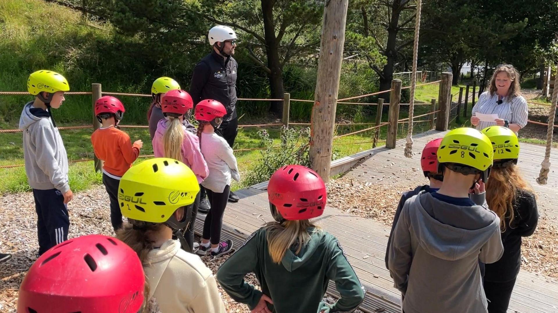 Children in active wear and yellow and red helmets watch a speech by the education minister, Daphne Caine. They are standing close to some rope swings.