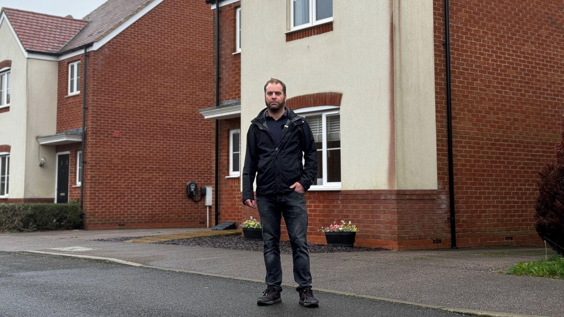 Dean Carpenter standing in front of his new build home with a large dark red stain running down one corner of the walls