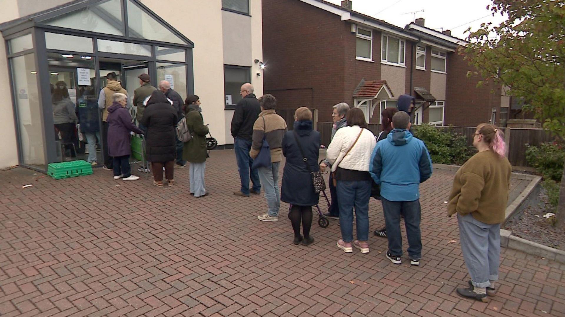 A queue of about a dozen people outside the door of a dental practice, with the queue then carrying on out of frame. People are dressed in coats and jumpers.