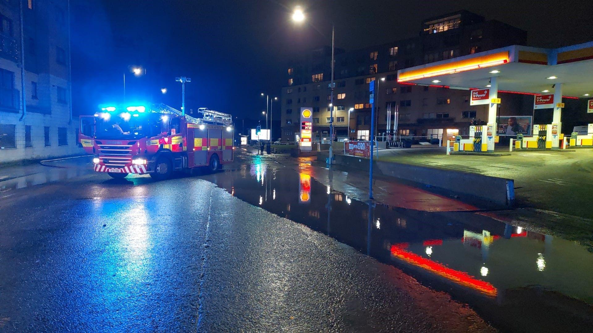 A fire engine with its blue lights on is parked on a road which is partially flooded next to a petrol station