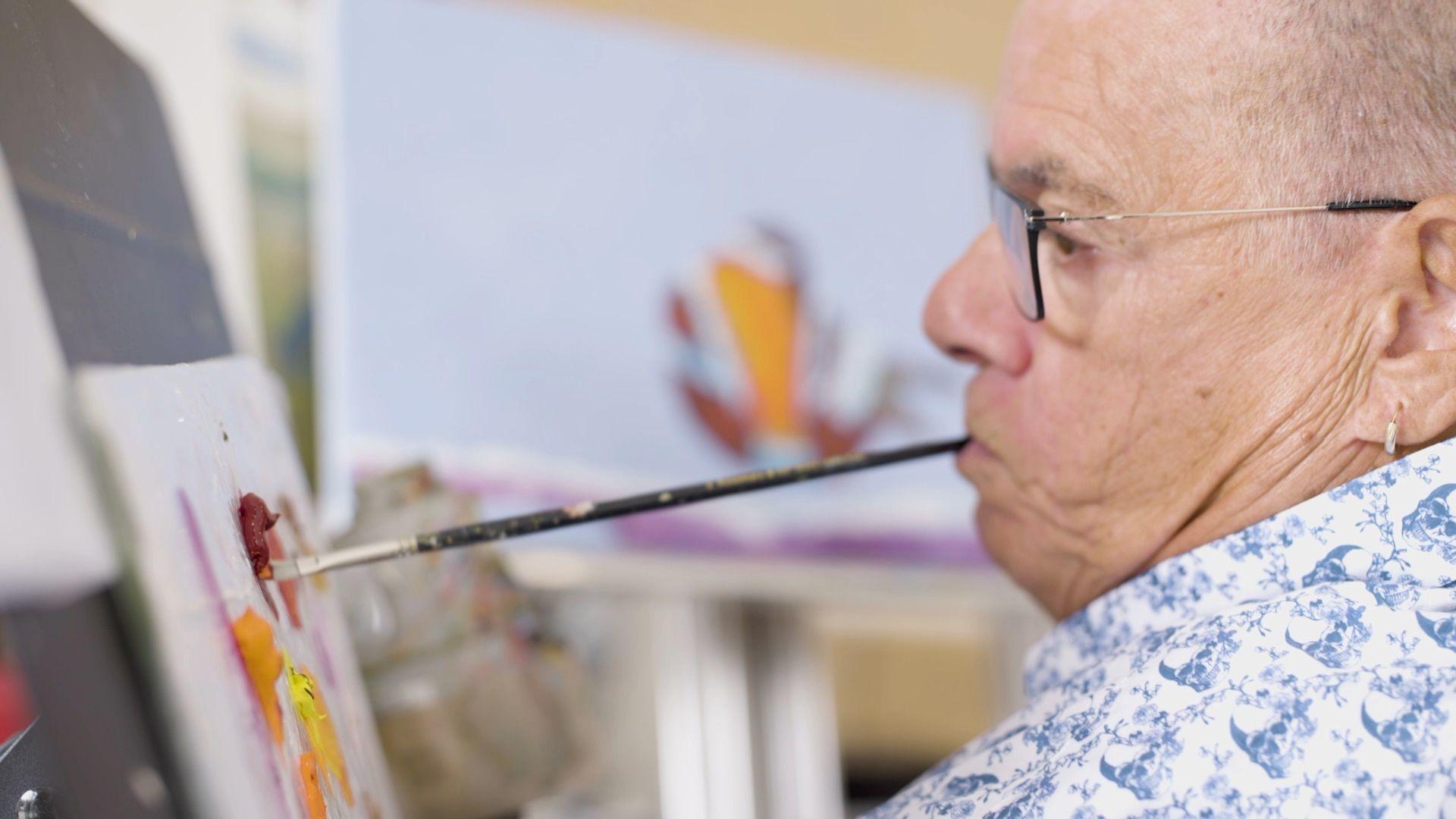 Rob Trent has short grey hair and wears thin black framed glasses, a silver hoop earring and a blue and white paisley print shirt. He is holding a black paintbrush in his mouth and dipping the paintbrush in a red paint which is on a stand to his left.