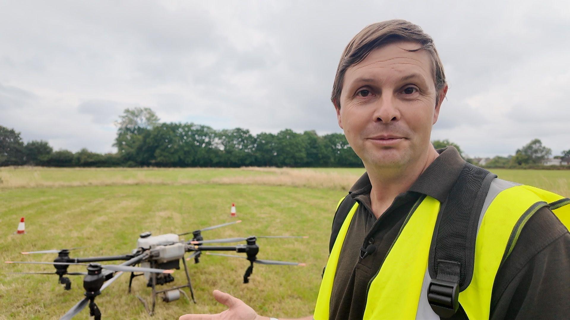A man wearing a yellow safety tabard stands next to a large farming drone ready to take off from a a field