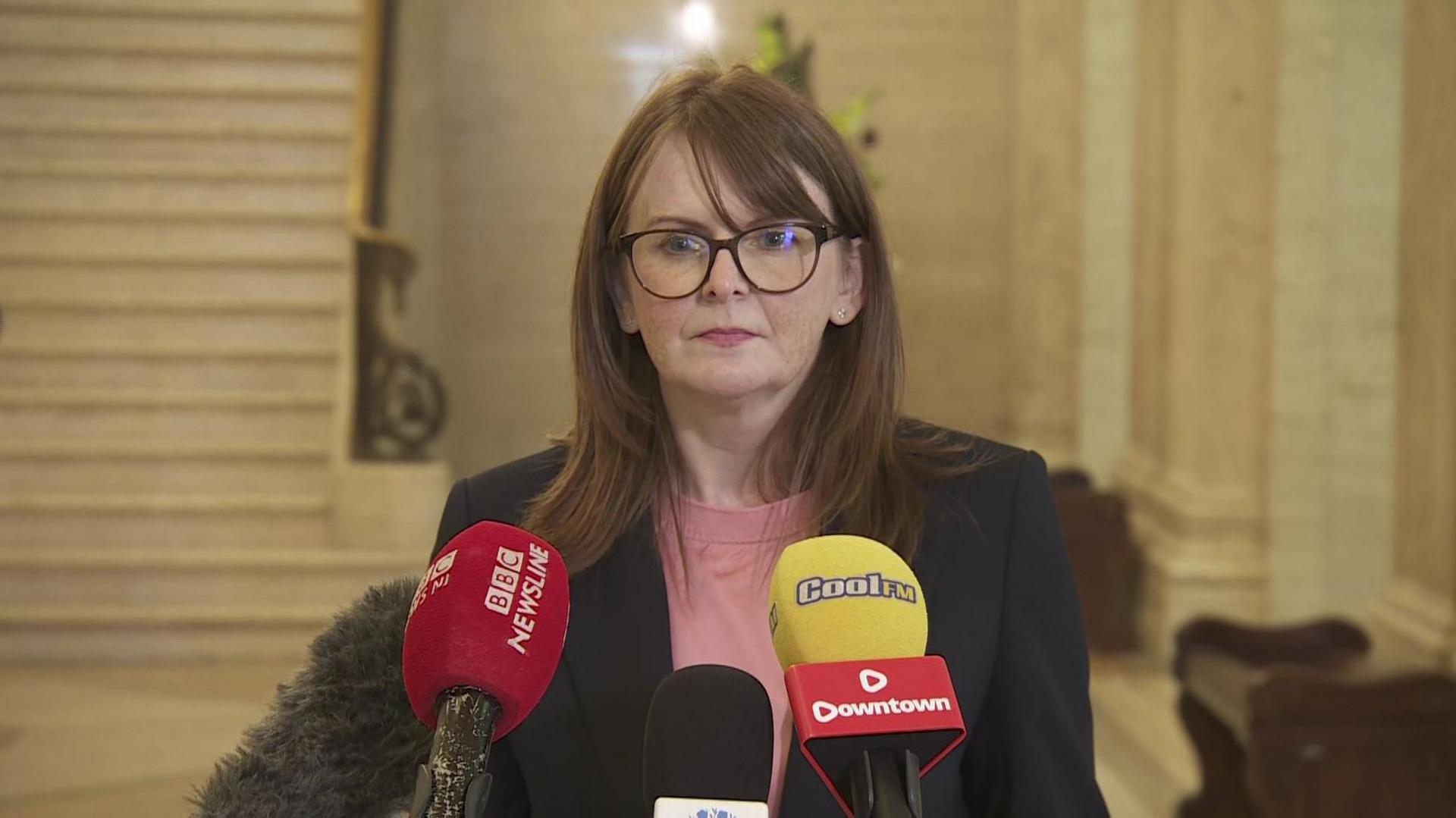 Finance Minister Caoimhe Archibald - A woman with long brown hair wearing large glasses with a dark frame. She is wearing a light pink top with a navy blazer, standing in front of press microphones. 