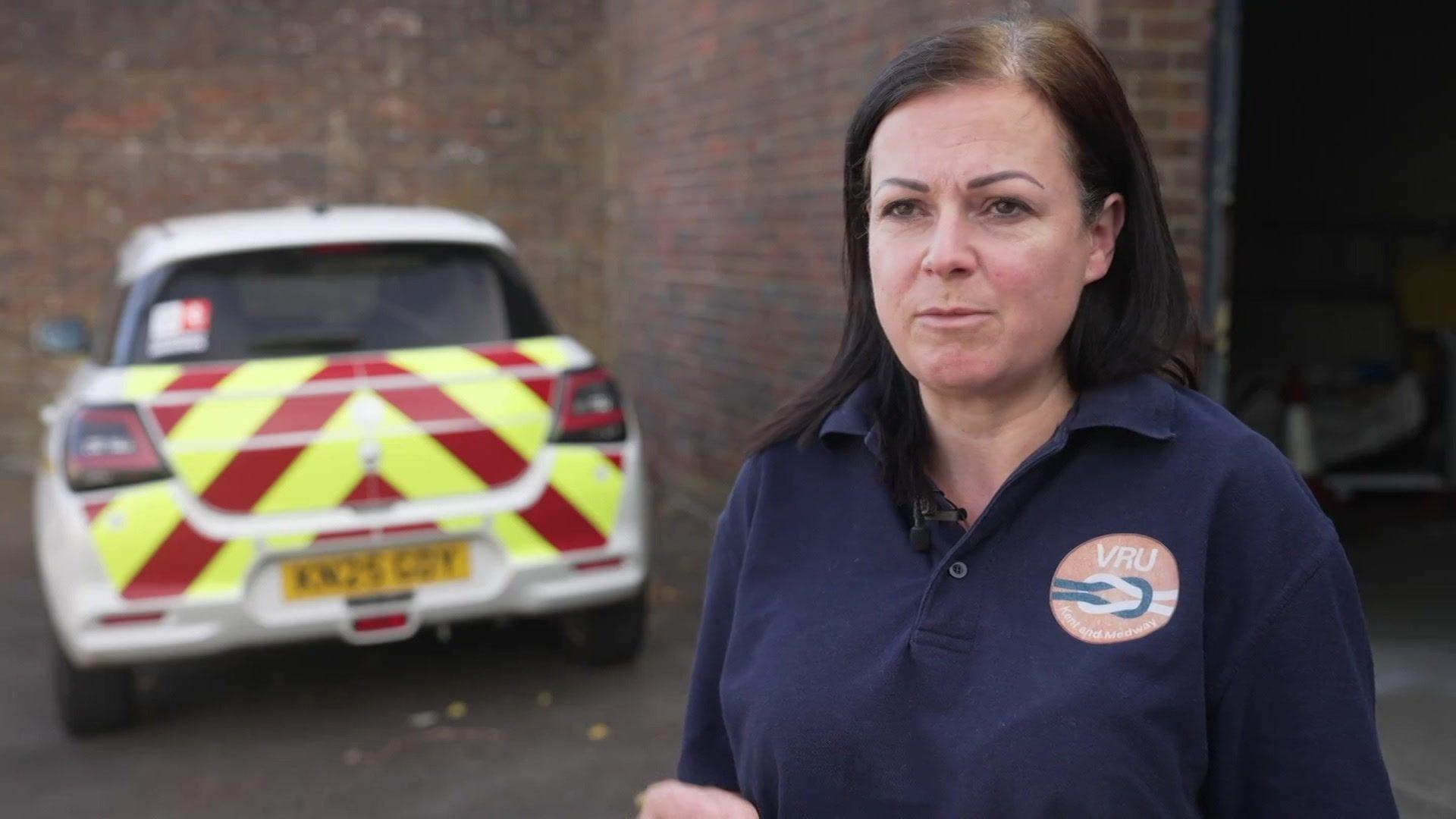 A woman dressed in a navy blue polo shirt with a logo on it with the letters "VRU" in white writing. She is stood in front of a car with a yellow and red hi-vis pattern on the back.